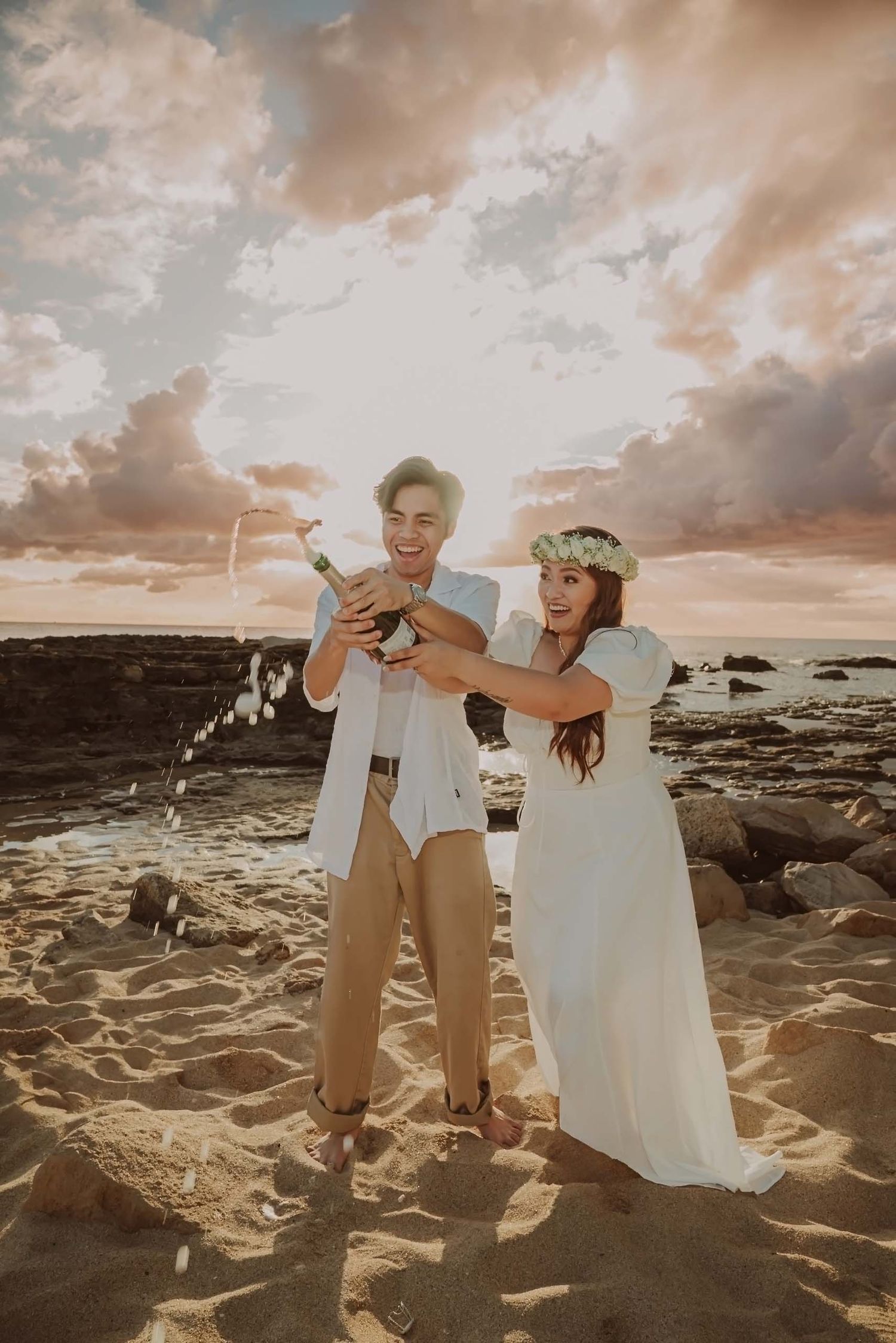 A couple shares a joyful moment on a rocky beach at sunset with dramatic clouds in the sky.