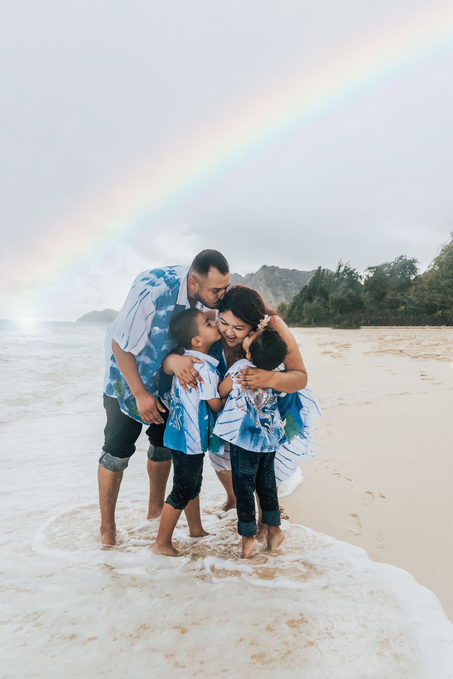 Family of four sharing a joyful embrace on a Hawaiian beach with a vibrant rainbow arching over lush mountains.