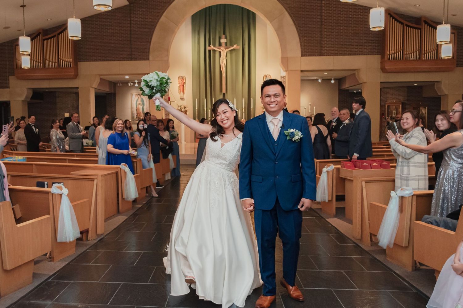 Newlywed couple walks down church aisle, bride in white gown holding bouquet, groom in blue suit, guests celebrating.