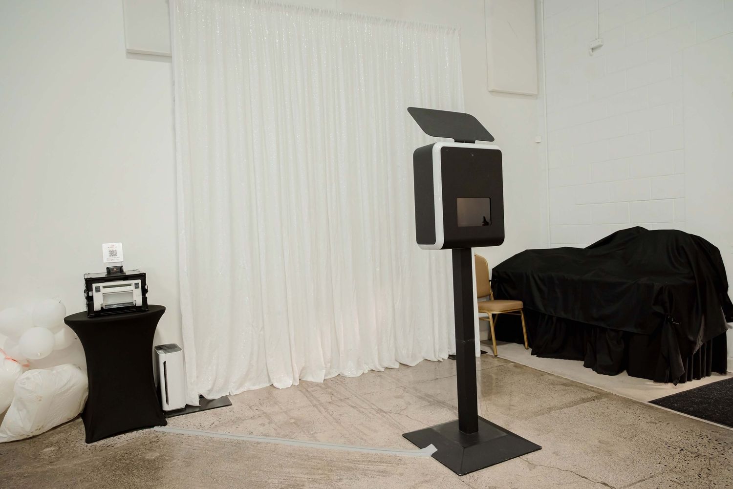 Modern photo booth on a stand in a white draped room with black covered furniture and tiled floors.