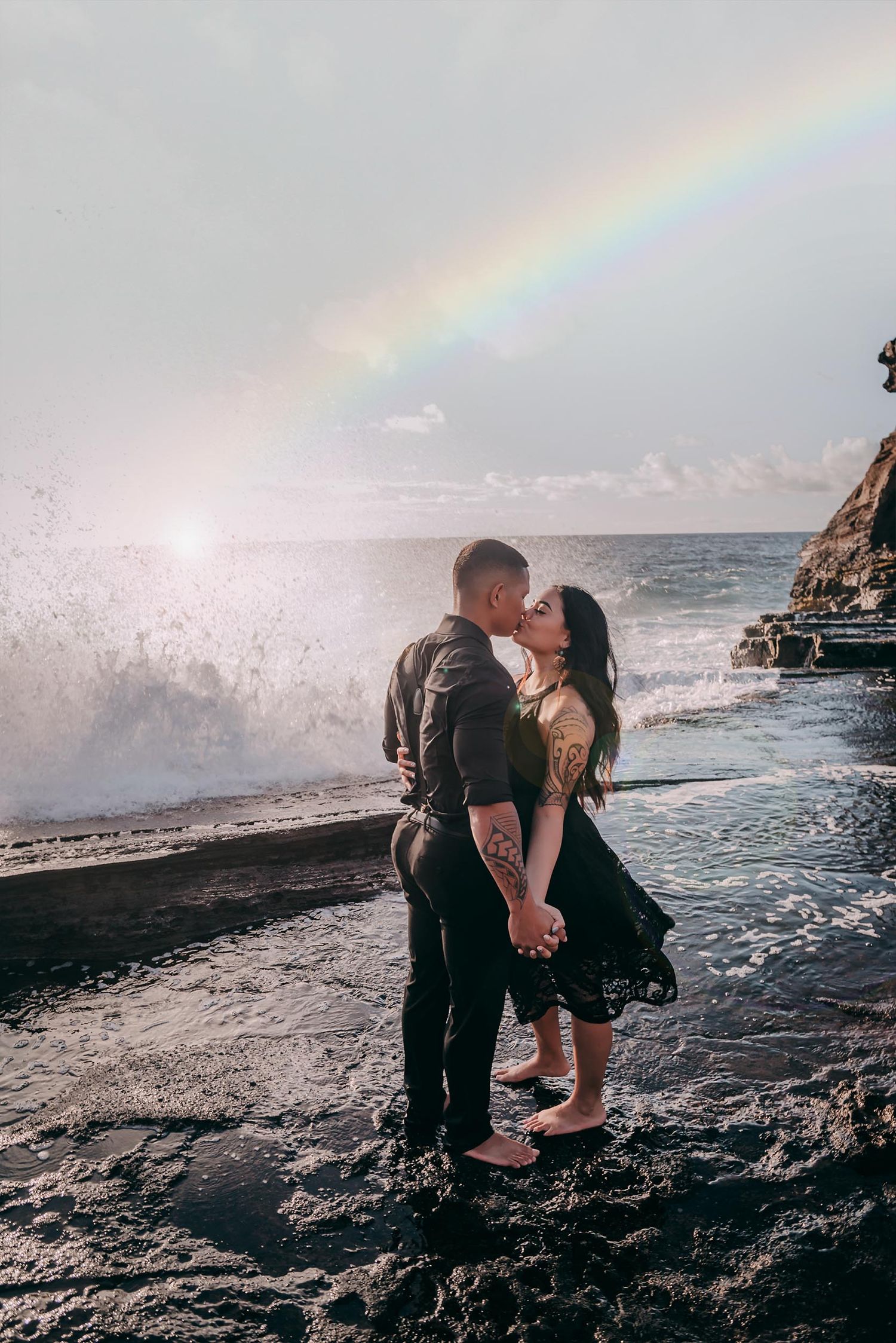 A couple shares a romantic moment on a rocky beach with a rainbow arcing over the ocean in the background.