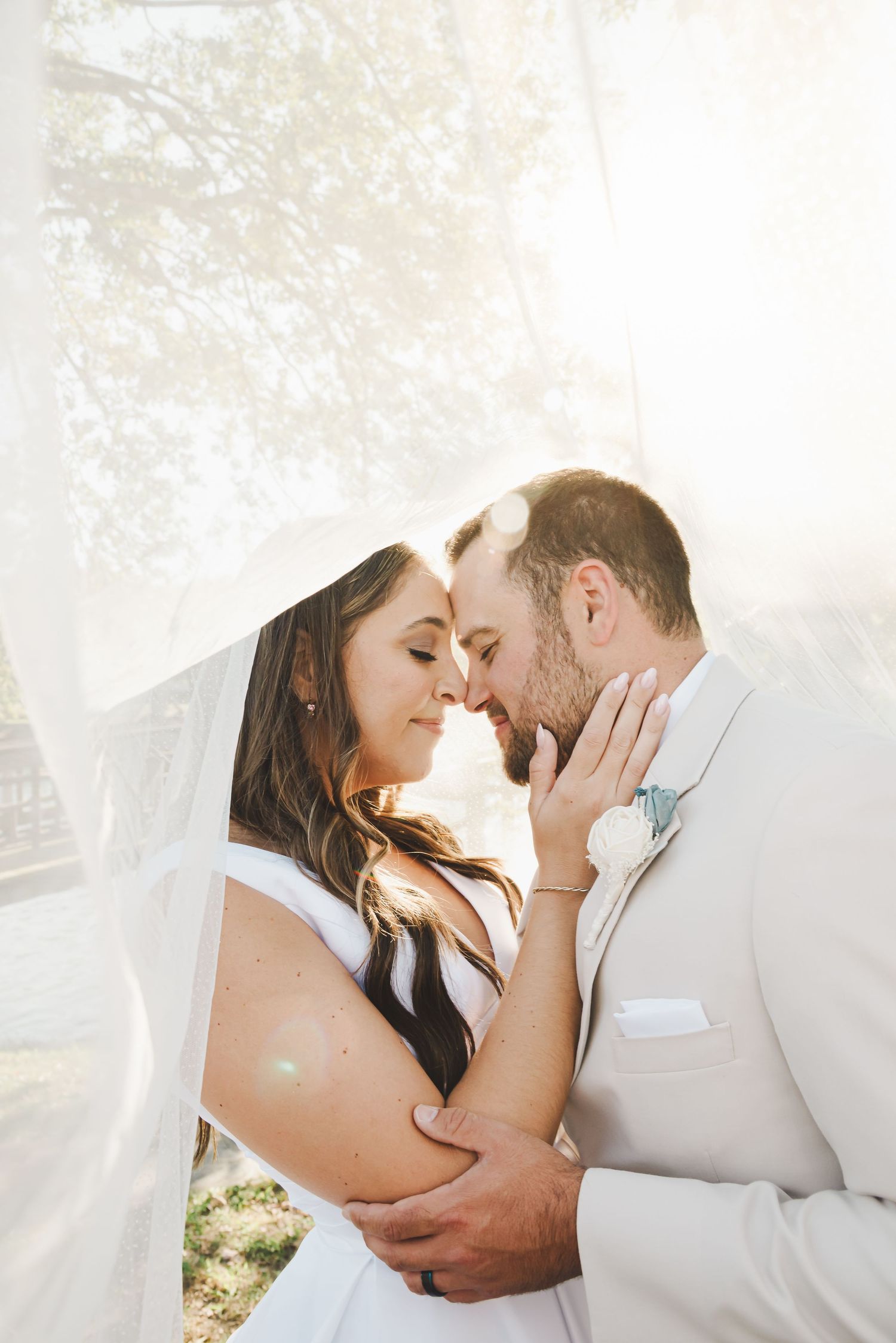 Romantic wedding couple sharing an intimate moment outdoors with sunlight filtering through a delicate veil.