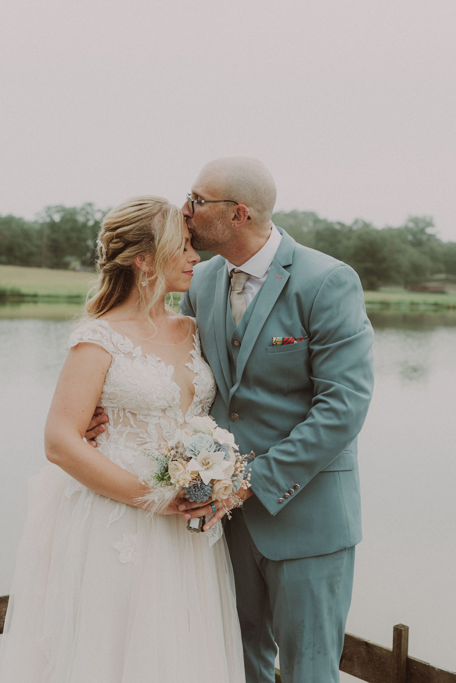 Romantic couple shares tender moments by a serene lakeside on their wedding day in matching blue and white attire.