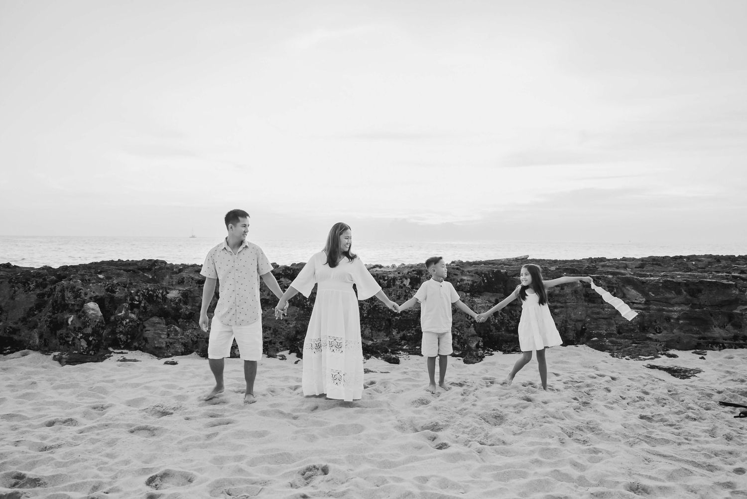 Black and white photo of a family of four holding hands on a sandy beach with rocky formations in the background.