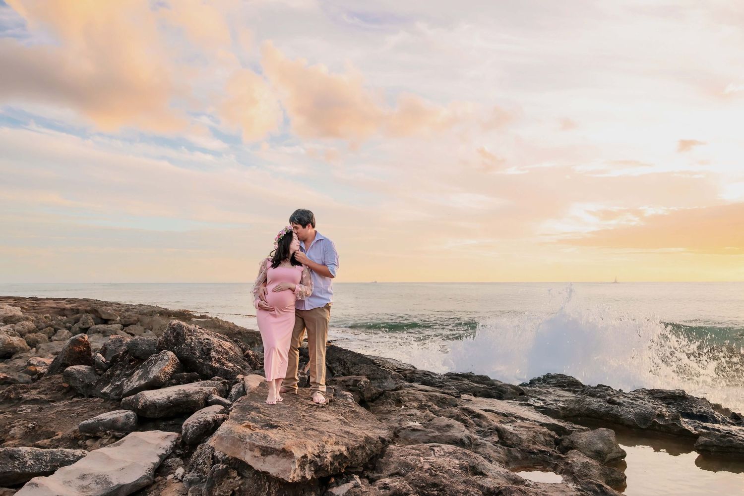 Couple embracing on rocky shoreline at sunset with ocean waves crashing behind them during a maternity photo session.