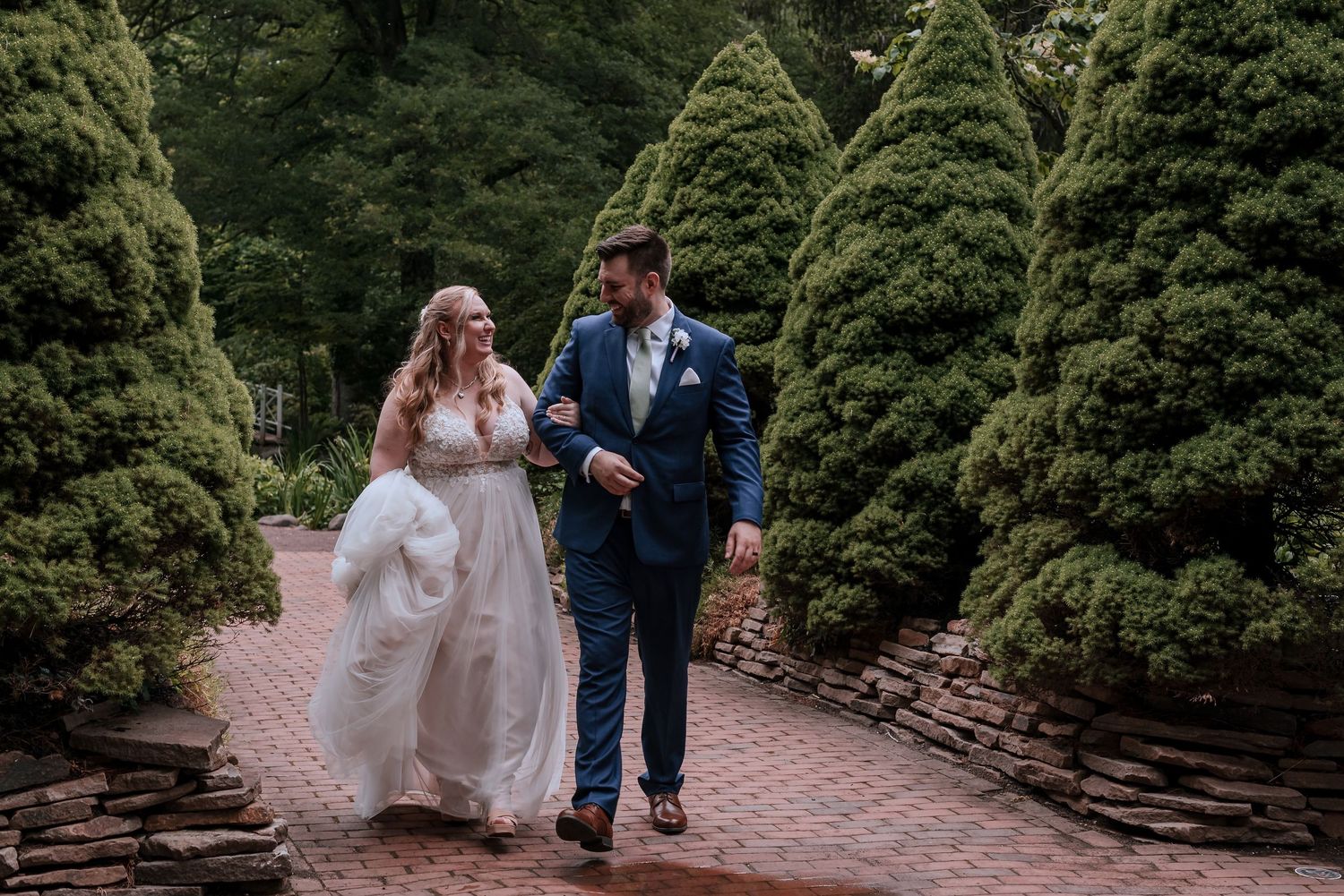 Bride and groom walking hand in hand through a lush garden pathway lined with evergreen trees on their wedding day.
