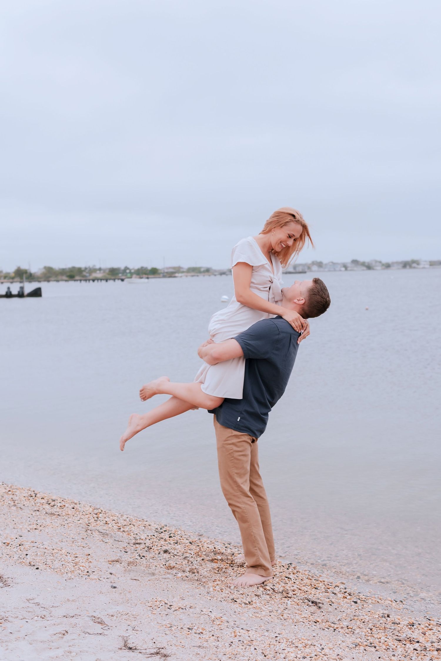 A romantic couple embraces on a beach shoreline with water and boats in the background.