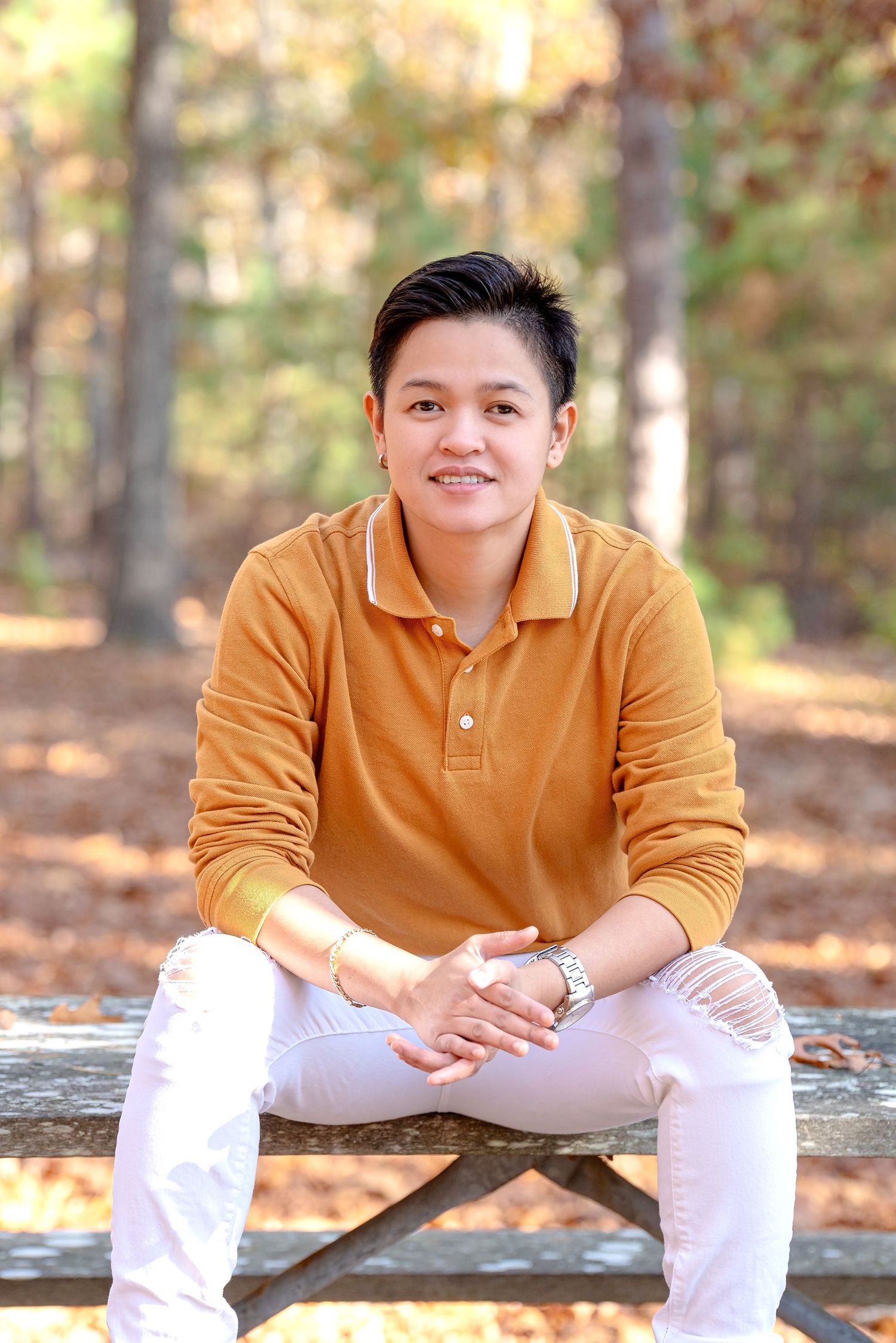 Person wearing a mustard yellow polo shirt sits casually on a wooden bench in an autumn forest setting.