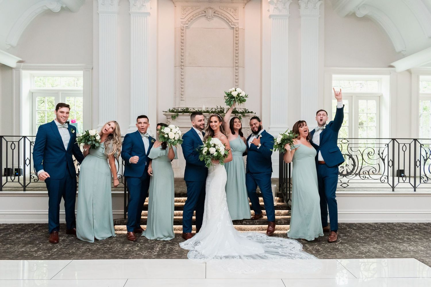 Wedding party in navy suits and mint bridesmaid dresses posing on elegant staircase in white venue.