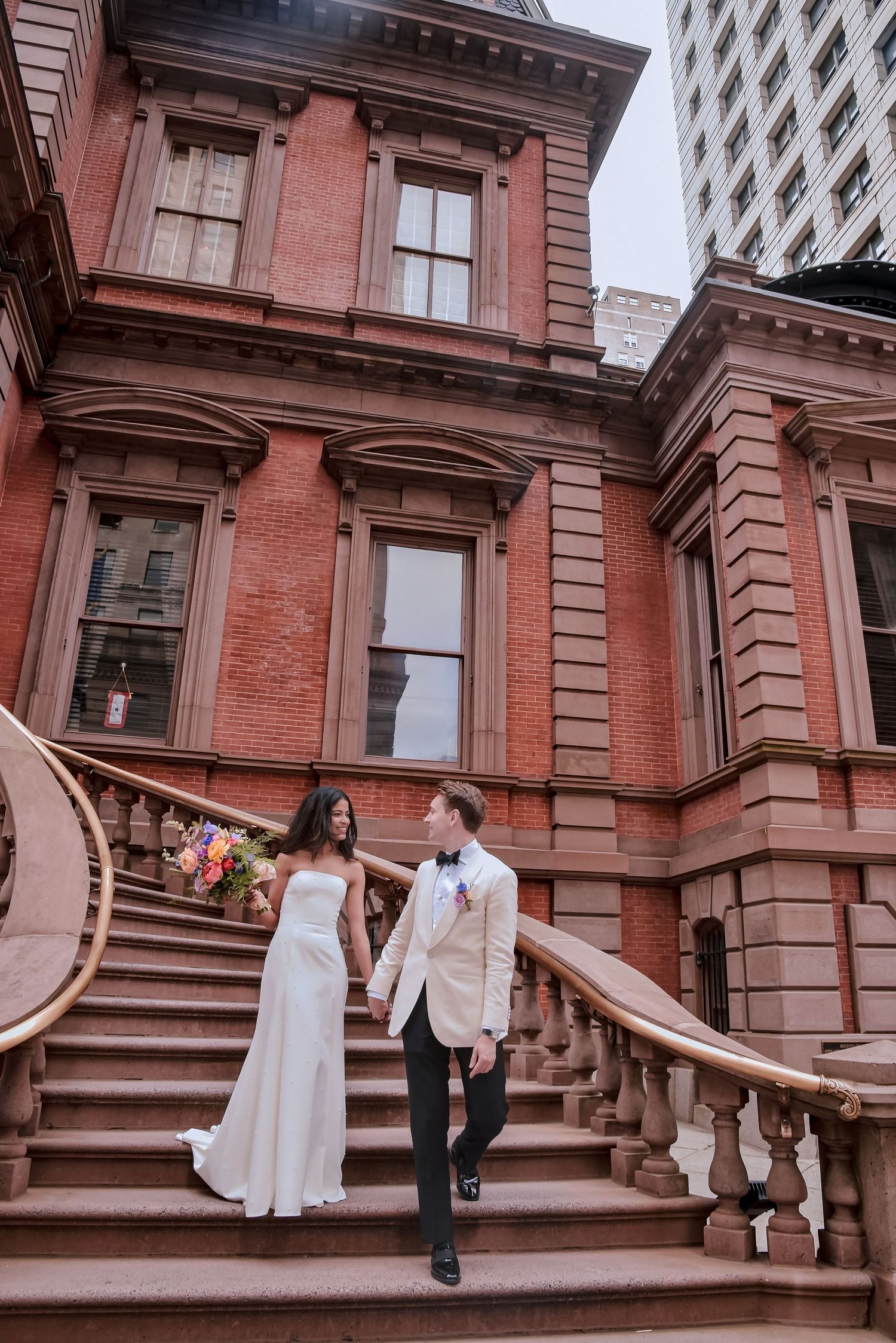 A couple poses on elegant red brick brownstone steps with ornate stone railings in a classic urban setting.