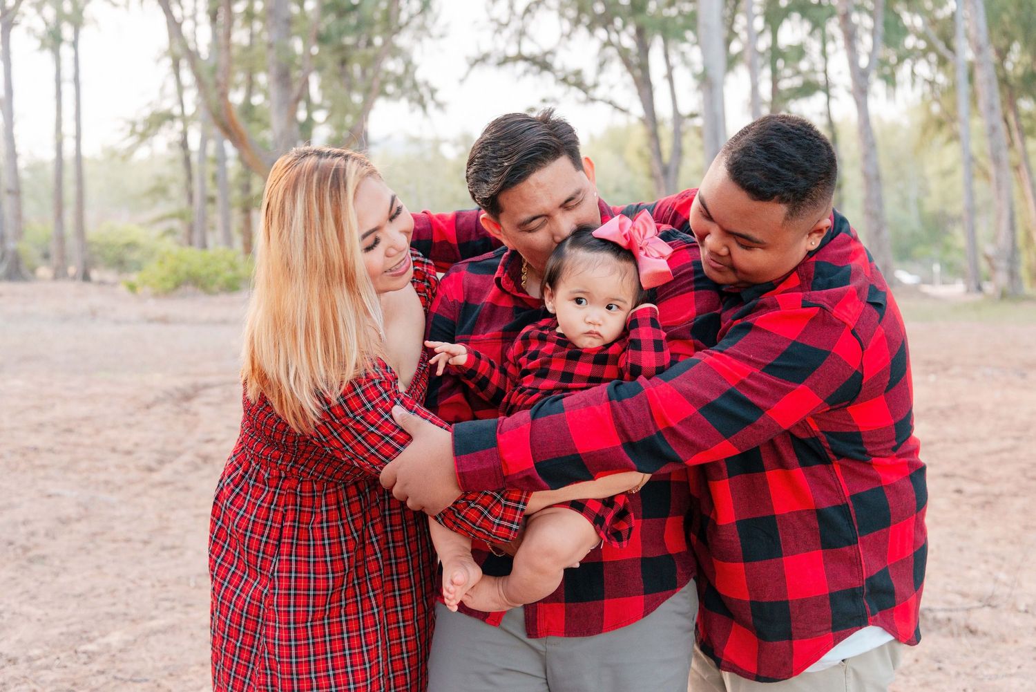 Family in matching red plaid shirts smiling and holding a baby outdoors among trees.