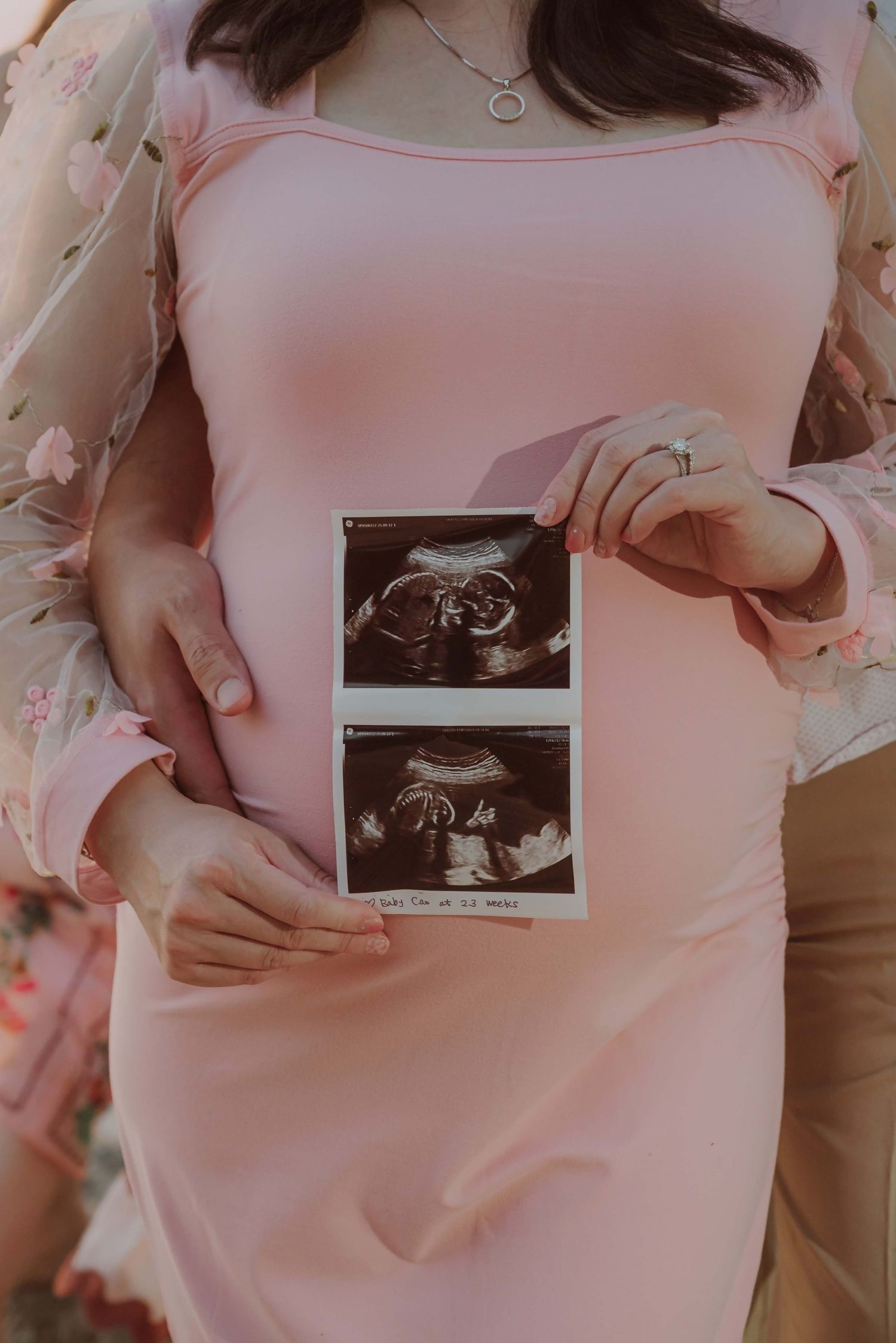 Pregnant woman in pink dress holding ultrasound photos while partner embraces her baby bump during maternity photo shoot.