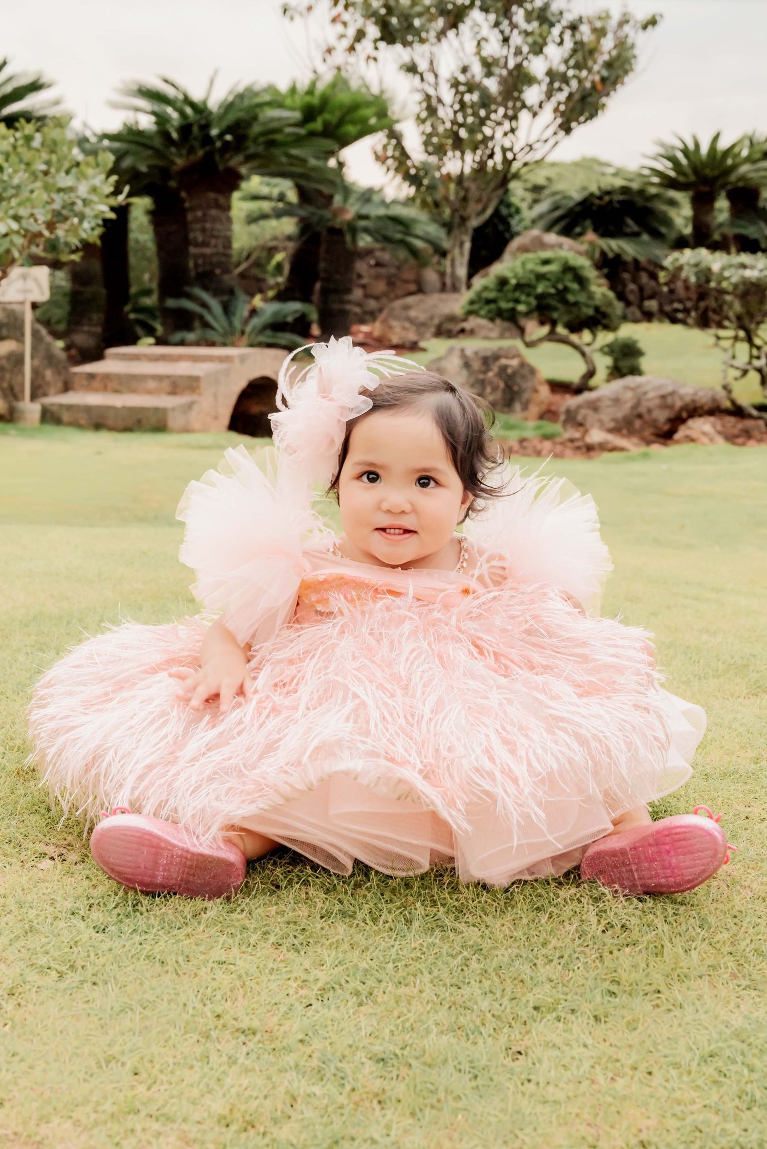 Adorable baby girl in a fluffy pink feather dress with tulle bow headband sitting on green grass in a garden.
