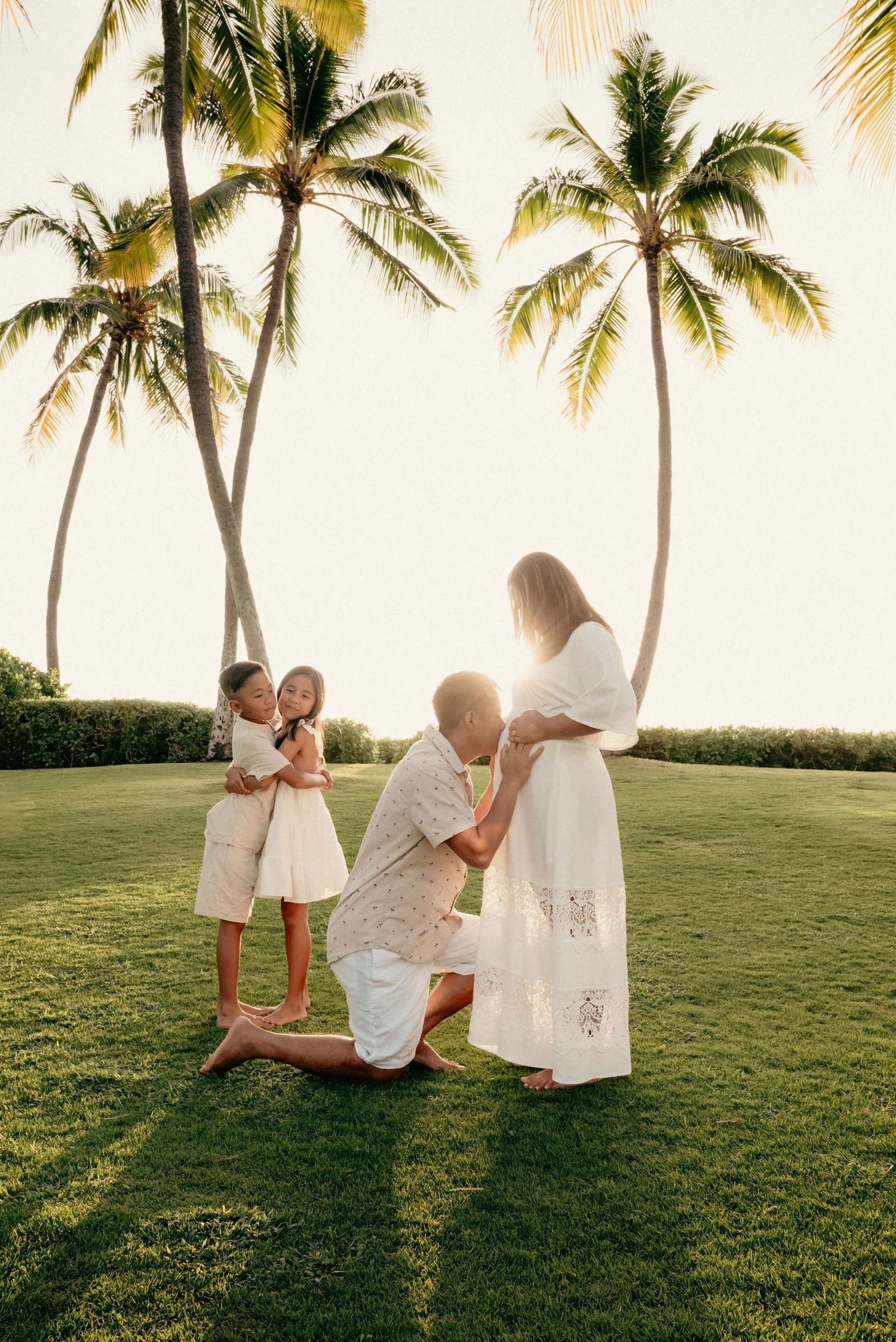 Family in white outfits poses on lush green lawn with tropical palm trees at golden sunset.