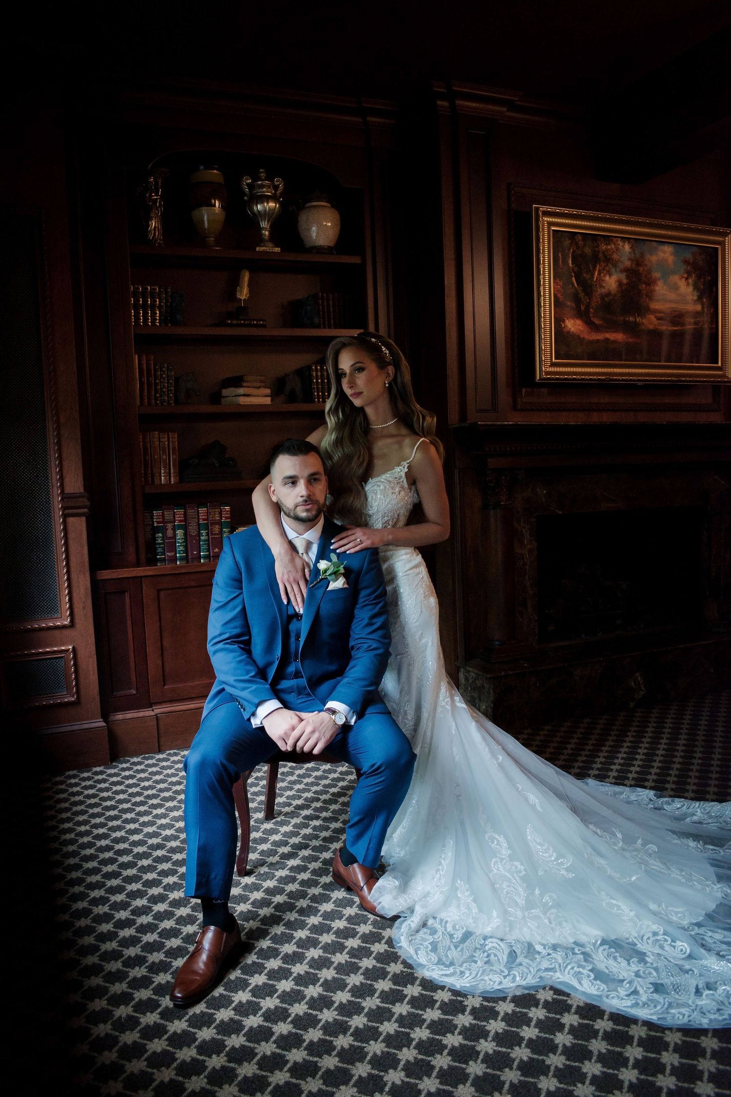 Elegant bride in lace gown stands beside groom in blue suit seated in chair inside a dark wood-paneled library.
