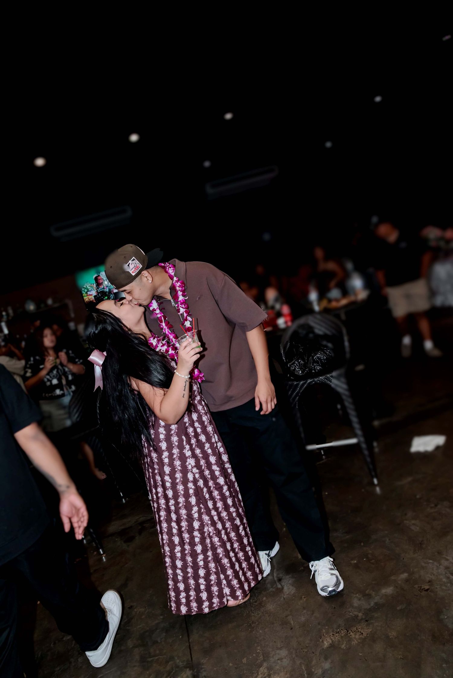 Couple sharing a romantic kiss at a nightclub event, woman in floral dress with lei, man in brown shirt and cap.