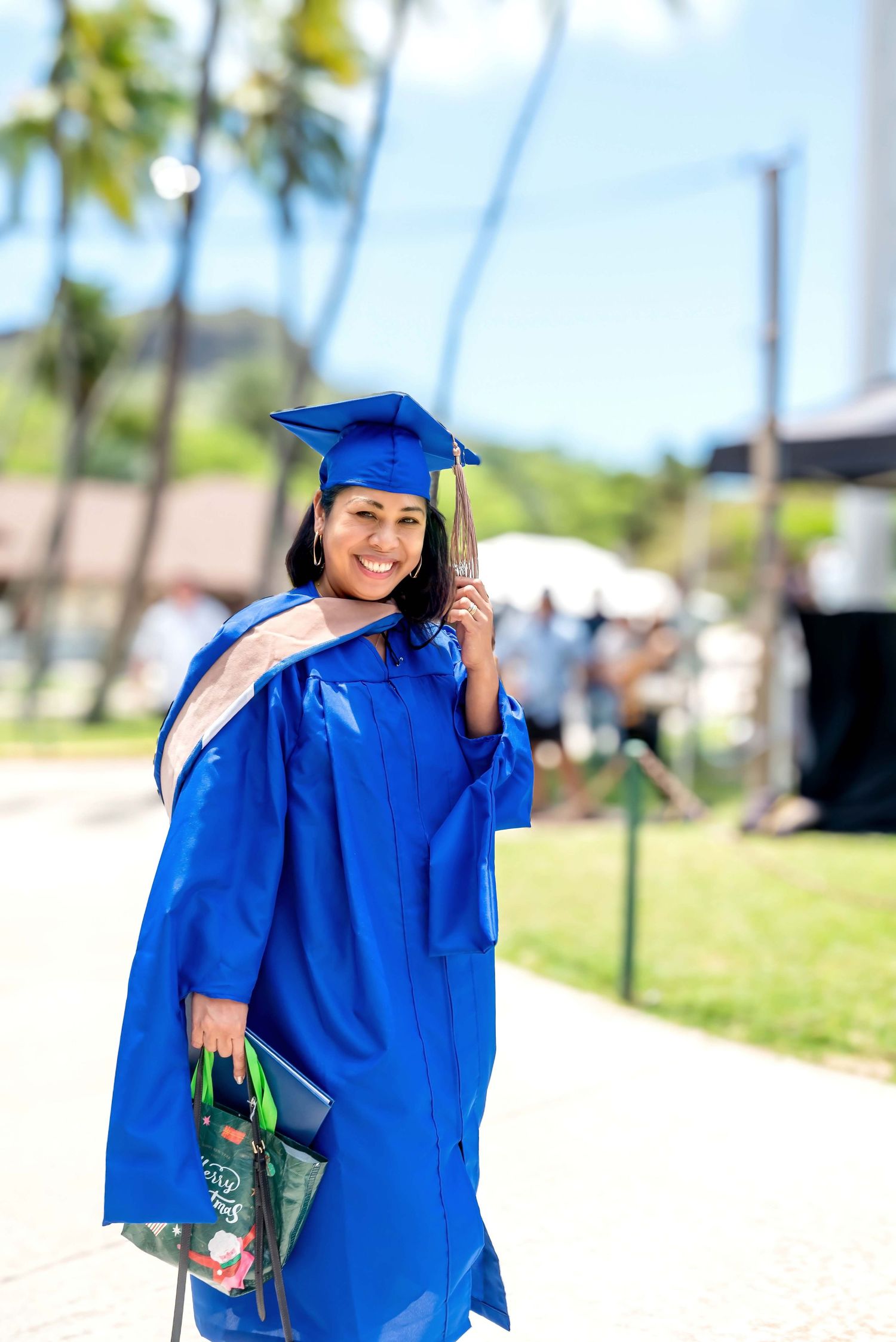 Smiling female graduate in blue cap and gown holding gifts outdoors near palm trees on a sunny day.