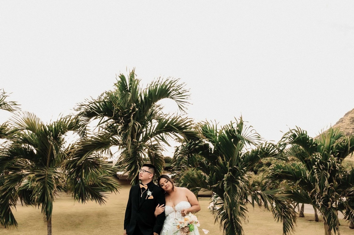 Bride and groom embrace beneath lush tropical palm trees on their wedding day outdoors.