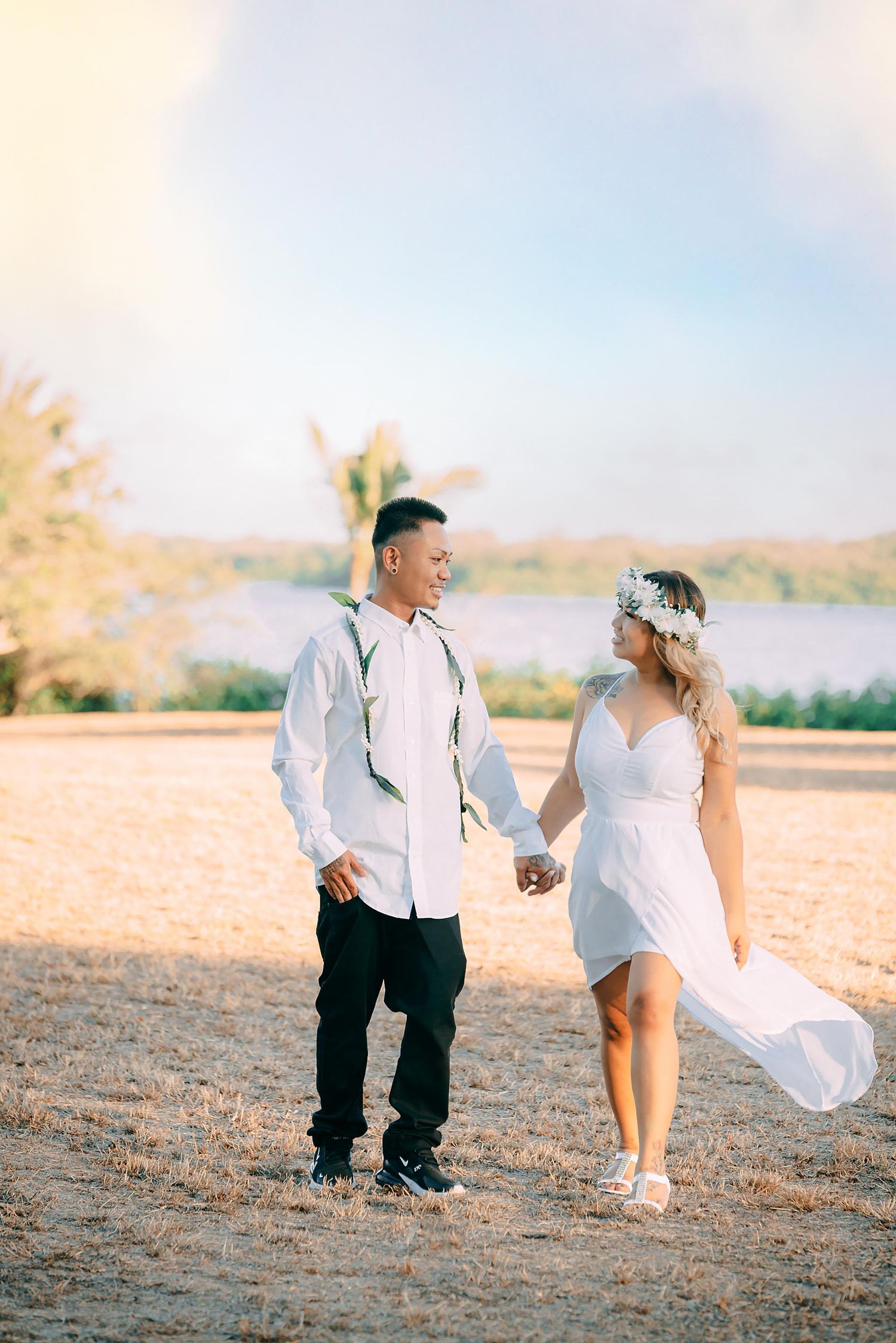 A couple share a romantic moment in a sunlit field with flowy white dress and casual attire against a scenic backdrop.