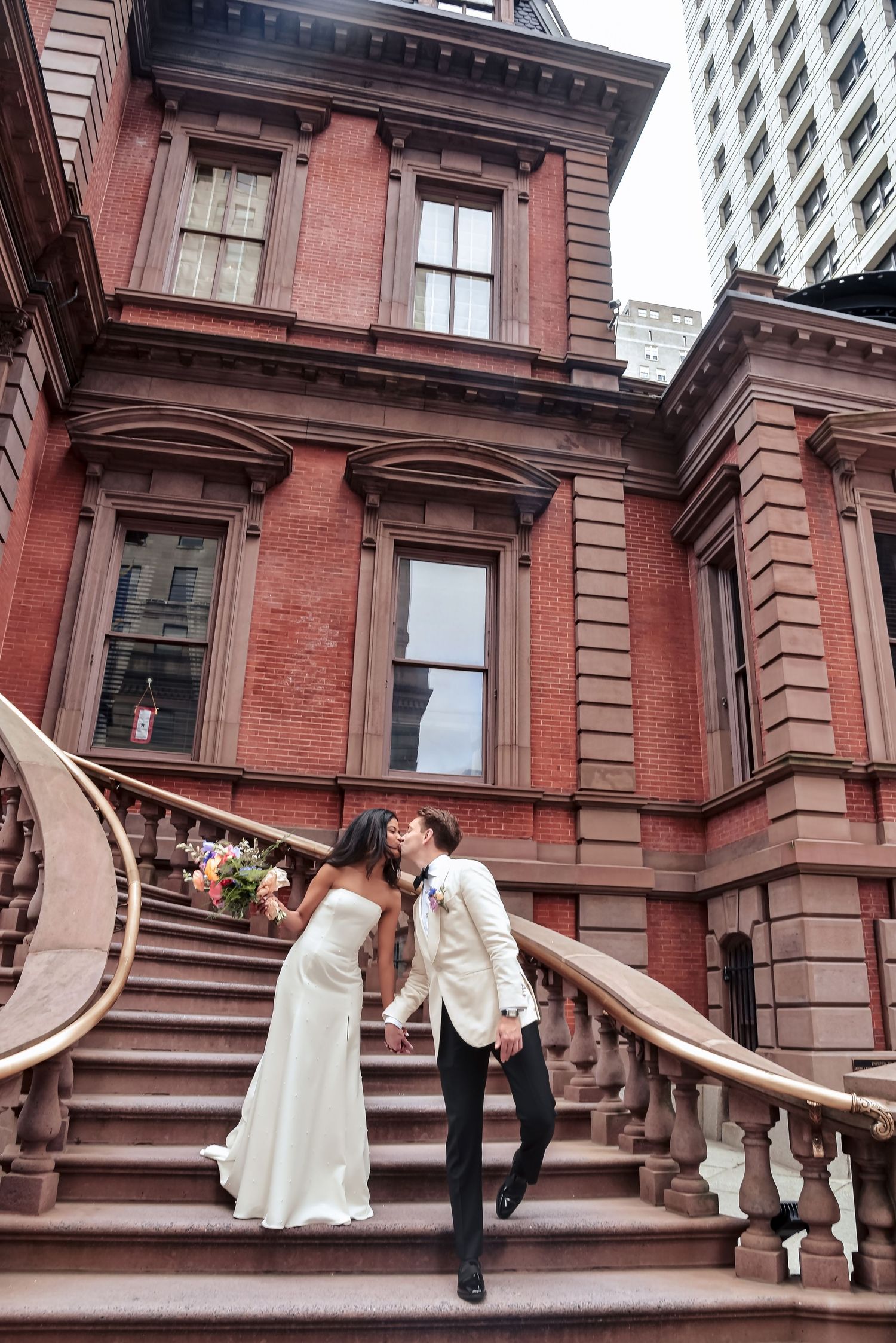 A romantic moment on the grand staircase of an ornate red brick Victorian mansion with decorative stone details.