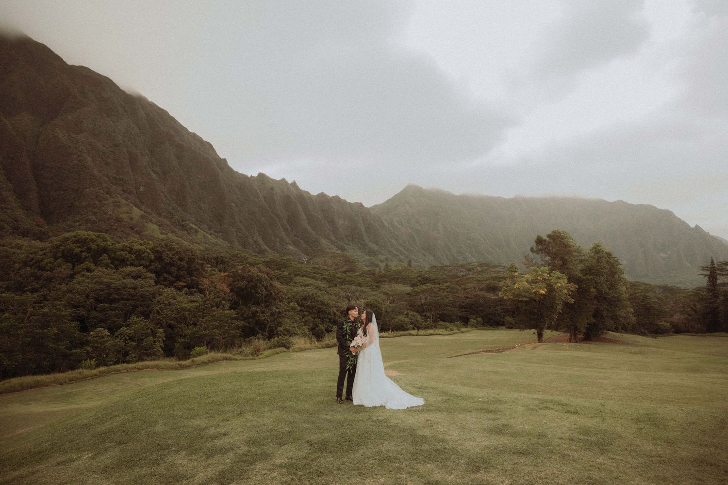 A couple shares a romantic moment in a misty mountain valley with dramatic green landscapes and cloudy skies.