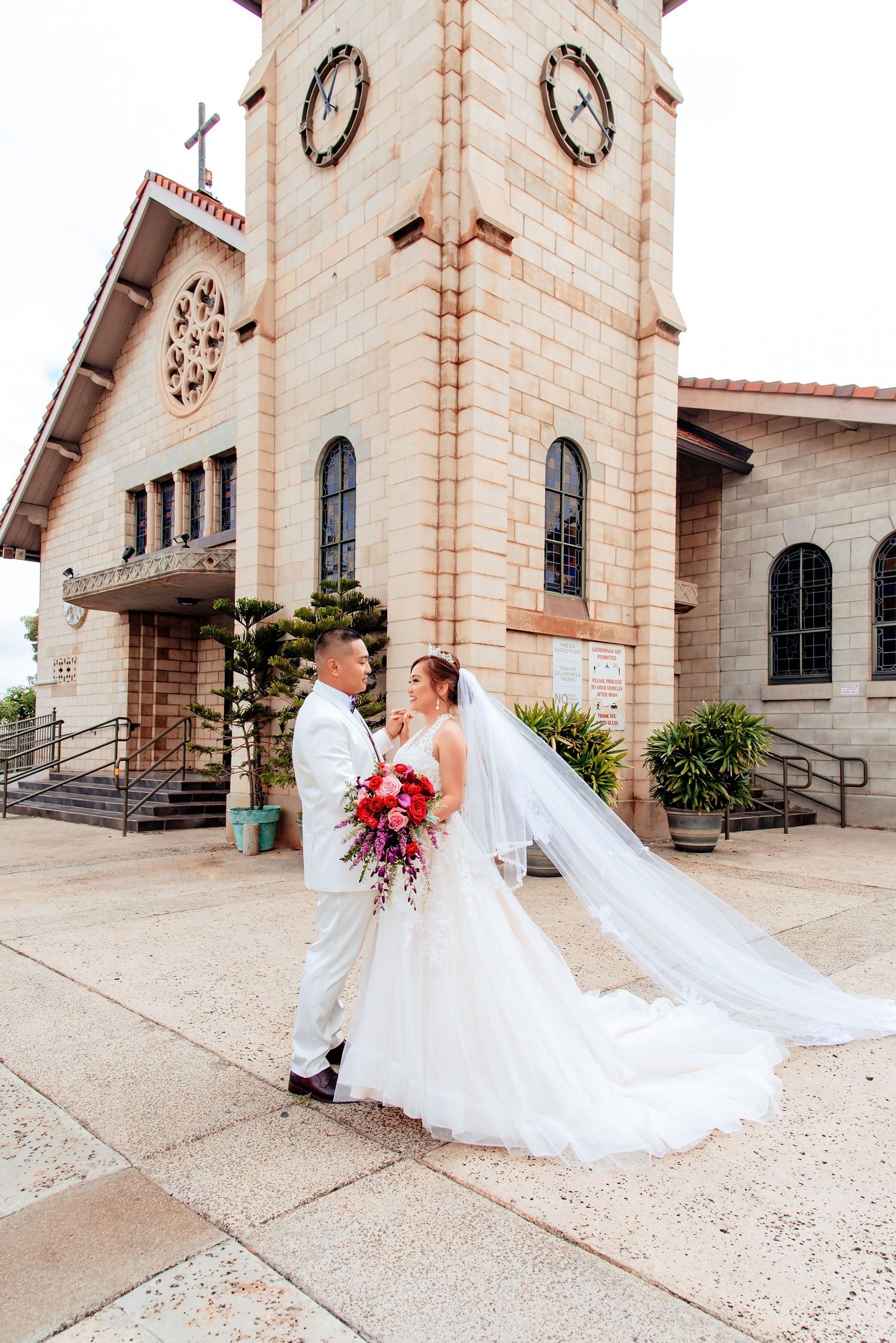 A stunning bride in a flowing white wedding gown with a dramatic cathedral veil poses outside a stone church.