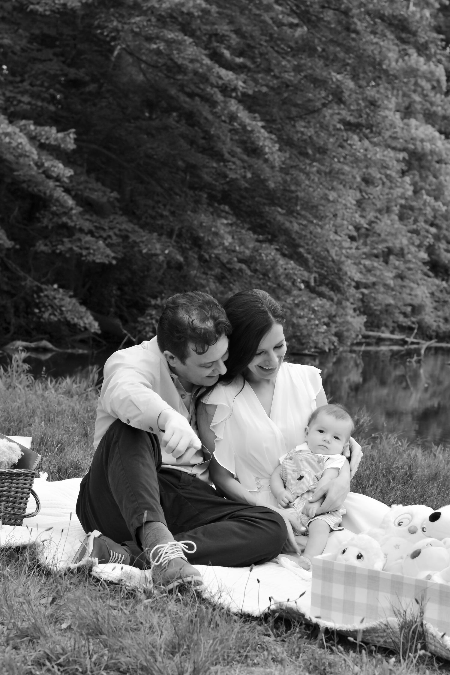 Black and white photo of a happy couple sitting on a picnic blanket outdoors, smiling at their newborn baby.