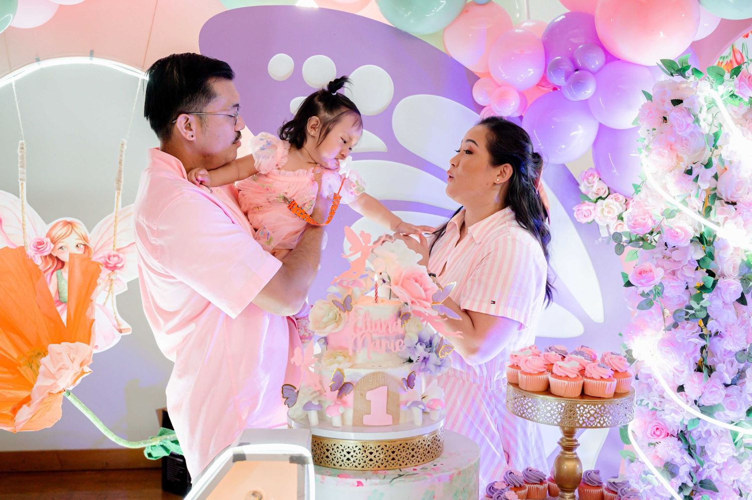 Family celebrating a baby girl's first birthday party with pastel balloons and colorful cake decorations.
