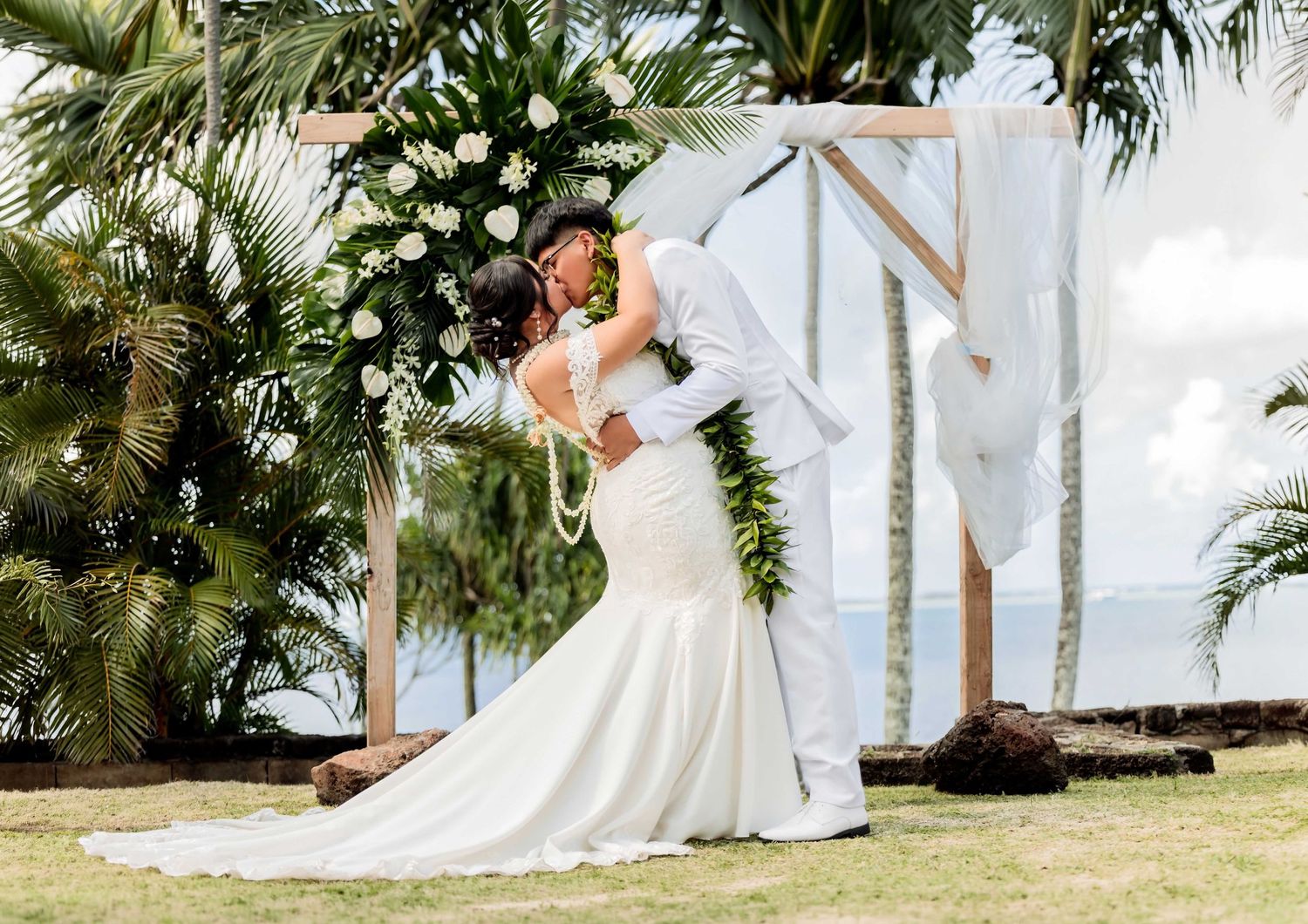 Bride and groom share a romantic kiss at outdoor wedding ceremony with floral arch and tropical palm trees backdrop.