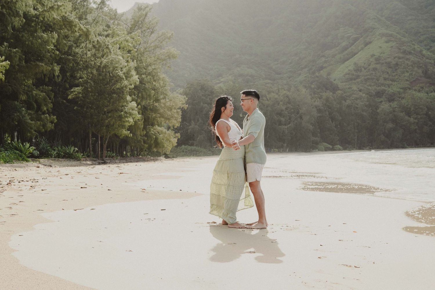 Couple standing barefoot on a sandy tropical beach, surrounded by lush green hills, sharing a romantic moment.