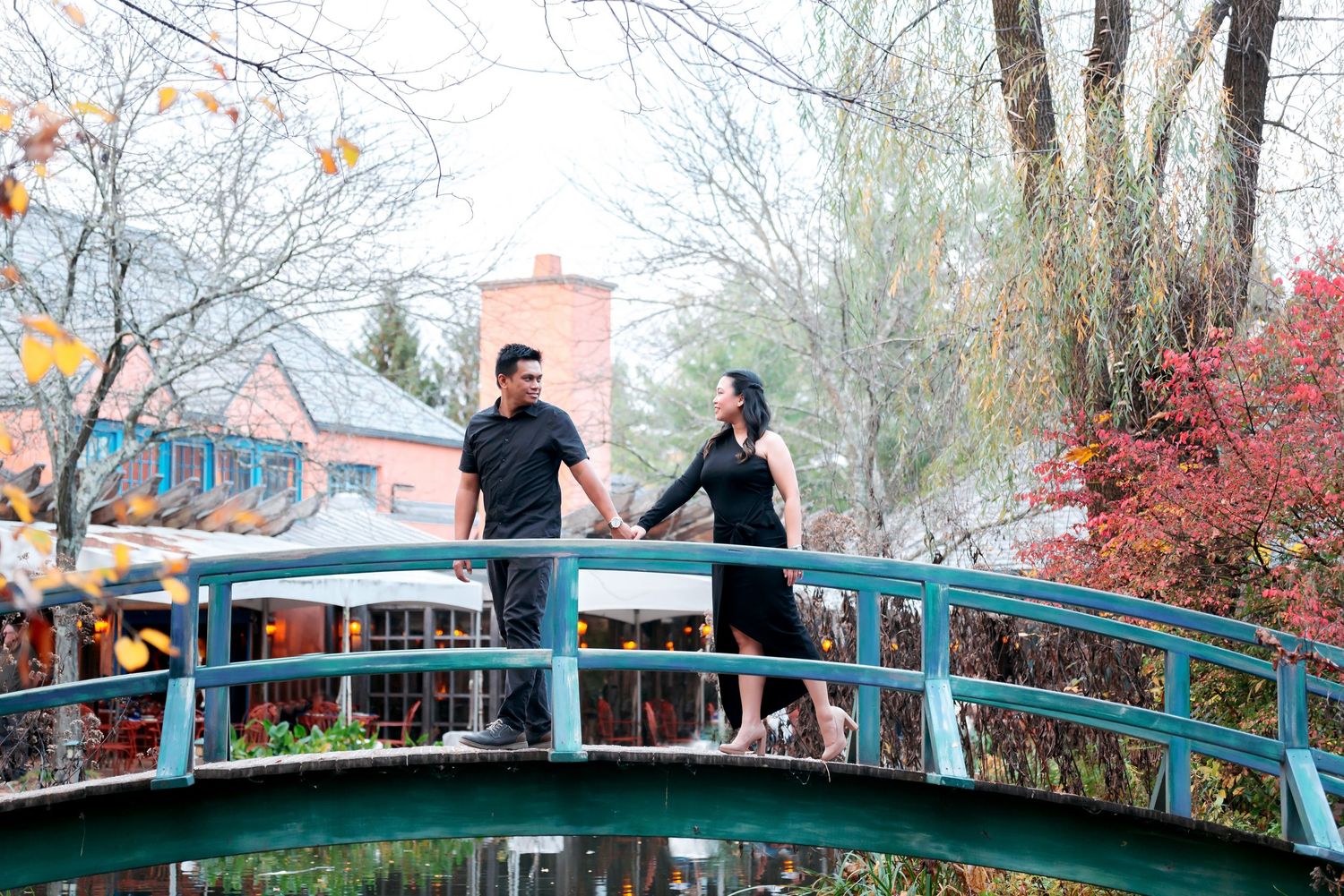 Couple holding hands on a turquoise arched bridge surrounded by autumn trees and colorful foliage.