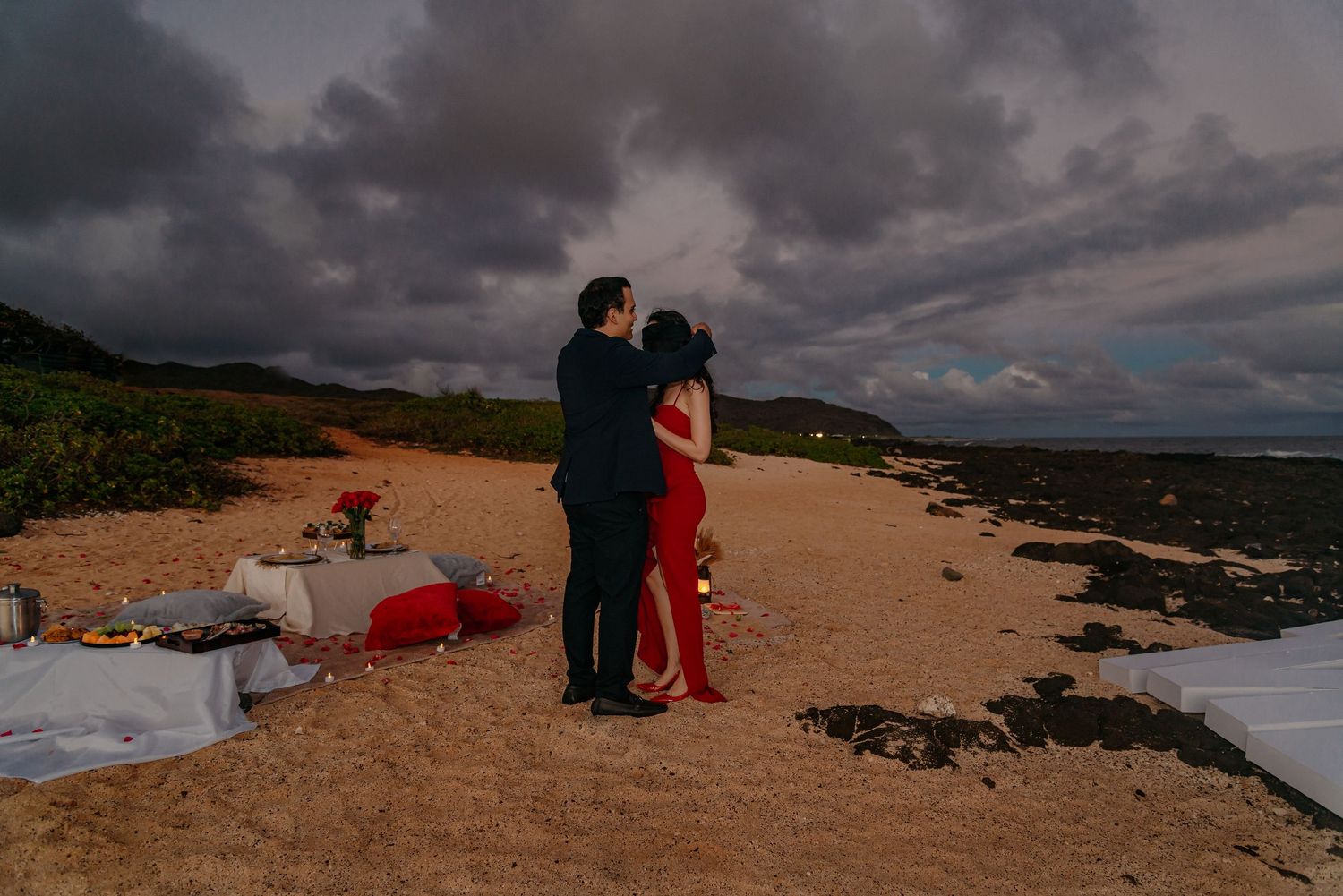 Couple embracing on a sandy beach at sunset with a romantic picnic setup and dramatic cloudy sky.