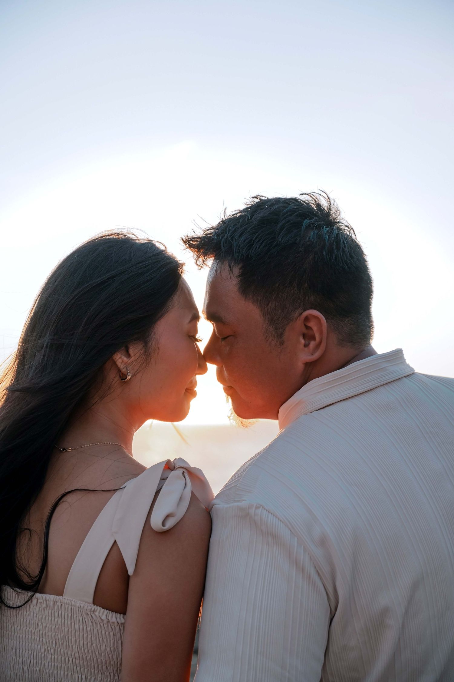 A romantic couple leans in close together at sunset, softly illuminated by warm golden light against a pale sky.