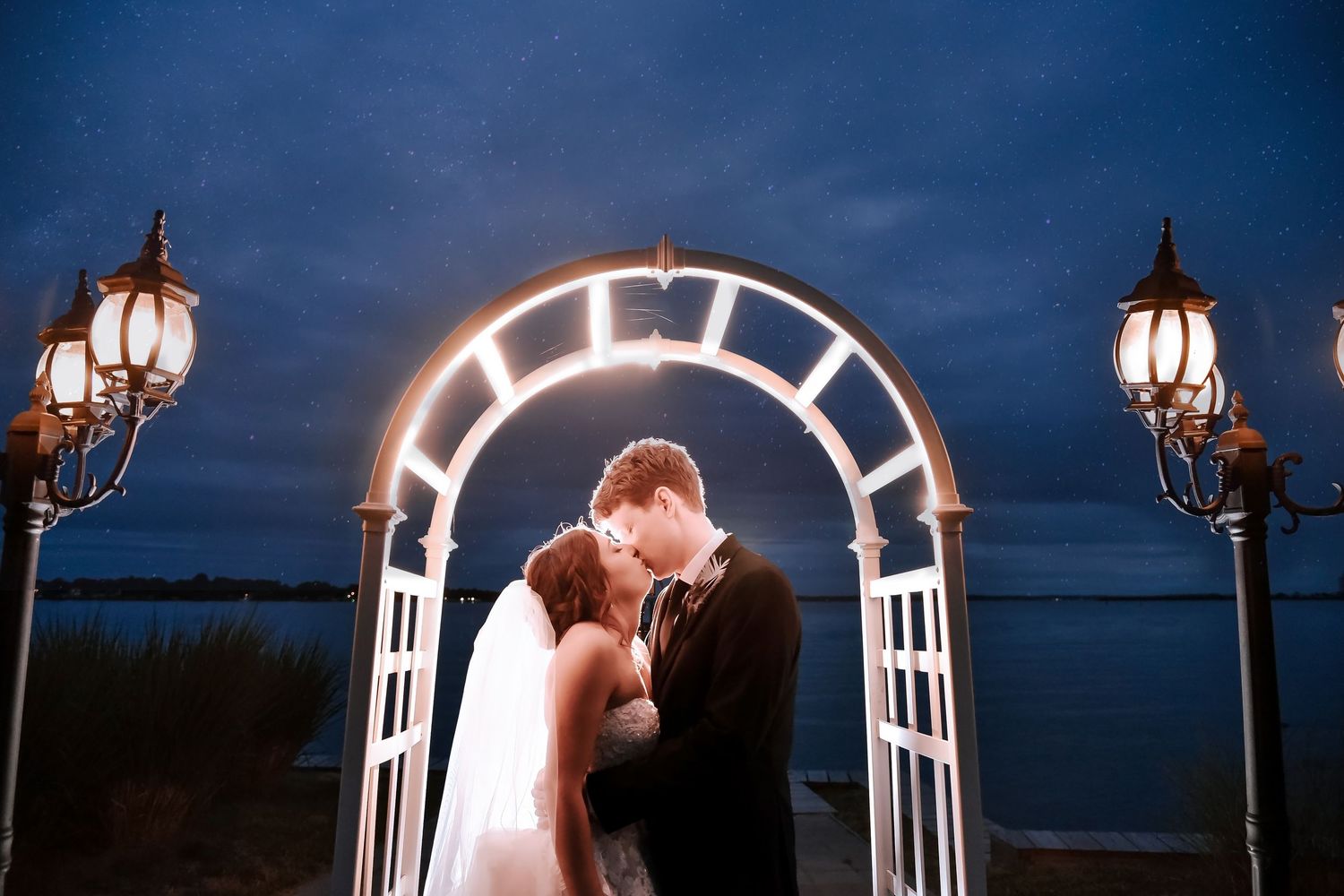 Bride and groom sharing a kiss under a glowing white arch with lanterns at dusk by the water.