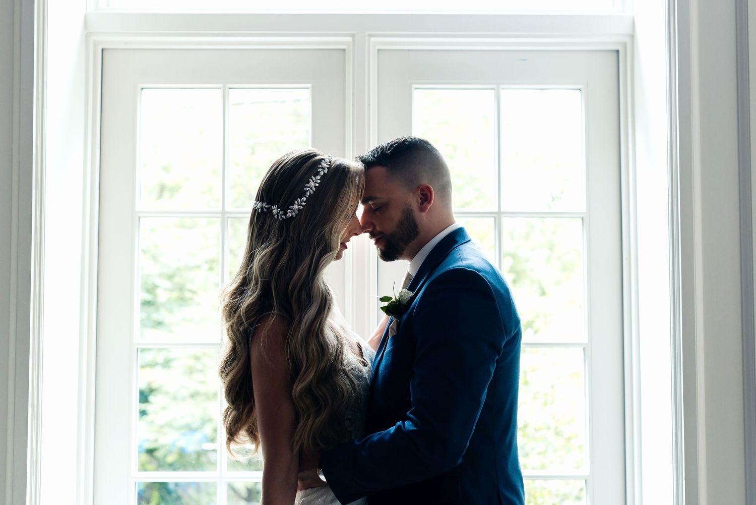 A bride and groom share an intimate moment, touching foreheads near a bright window on their wedding day.