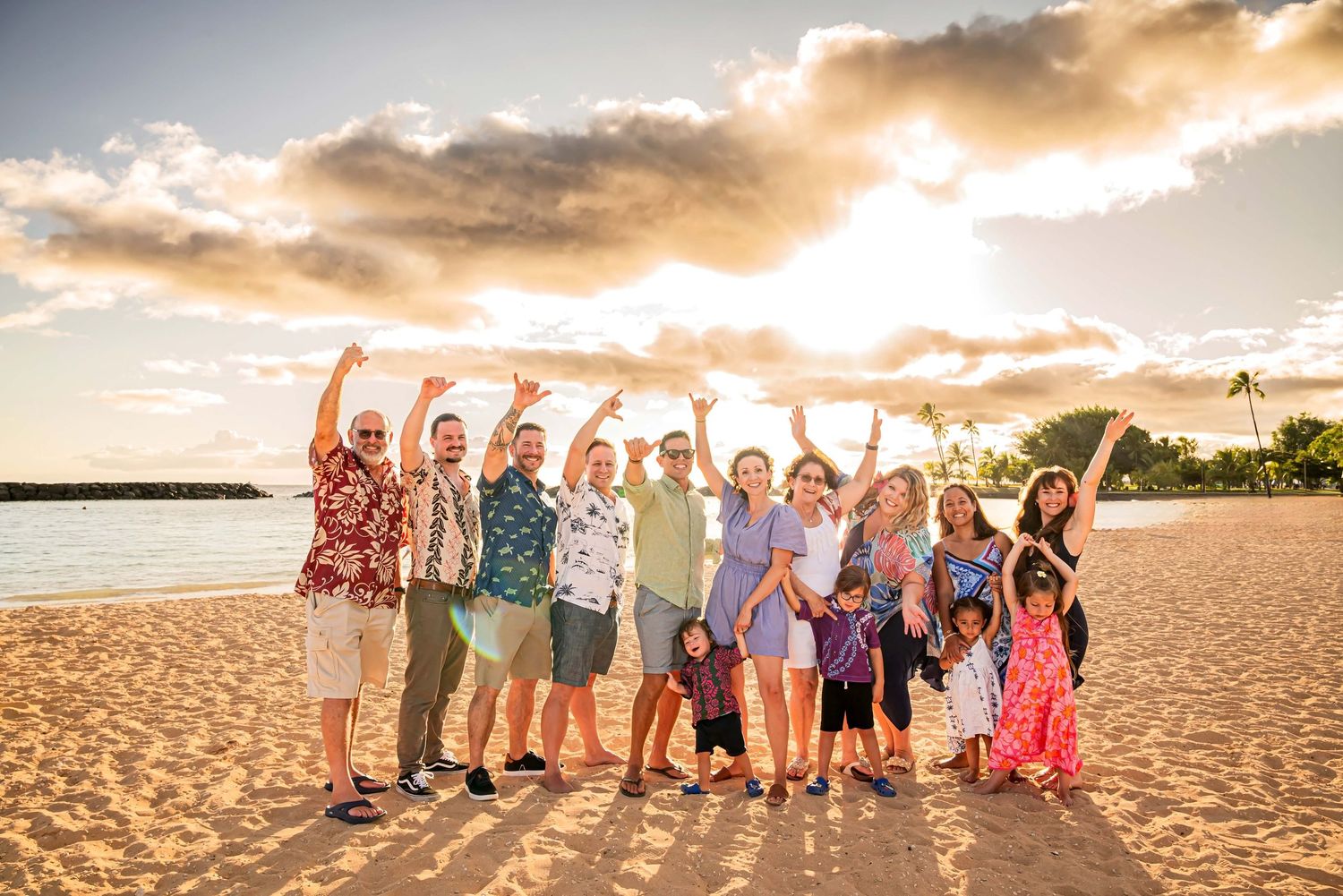Large family group celebrating on a sandy beach at sunset in Hawaii, with arms raised joyfully.