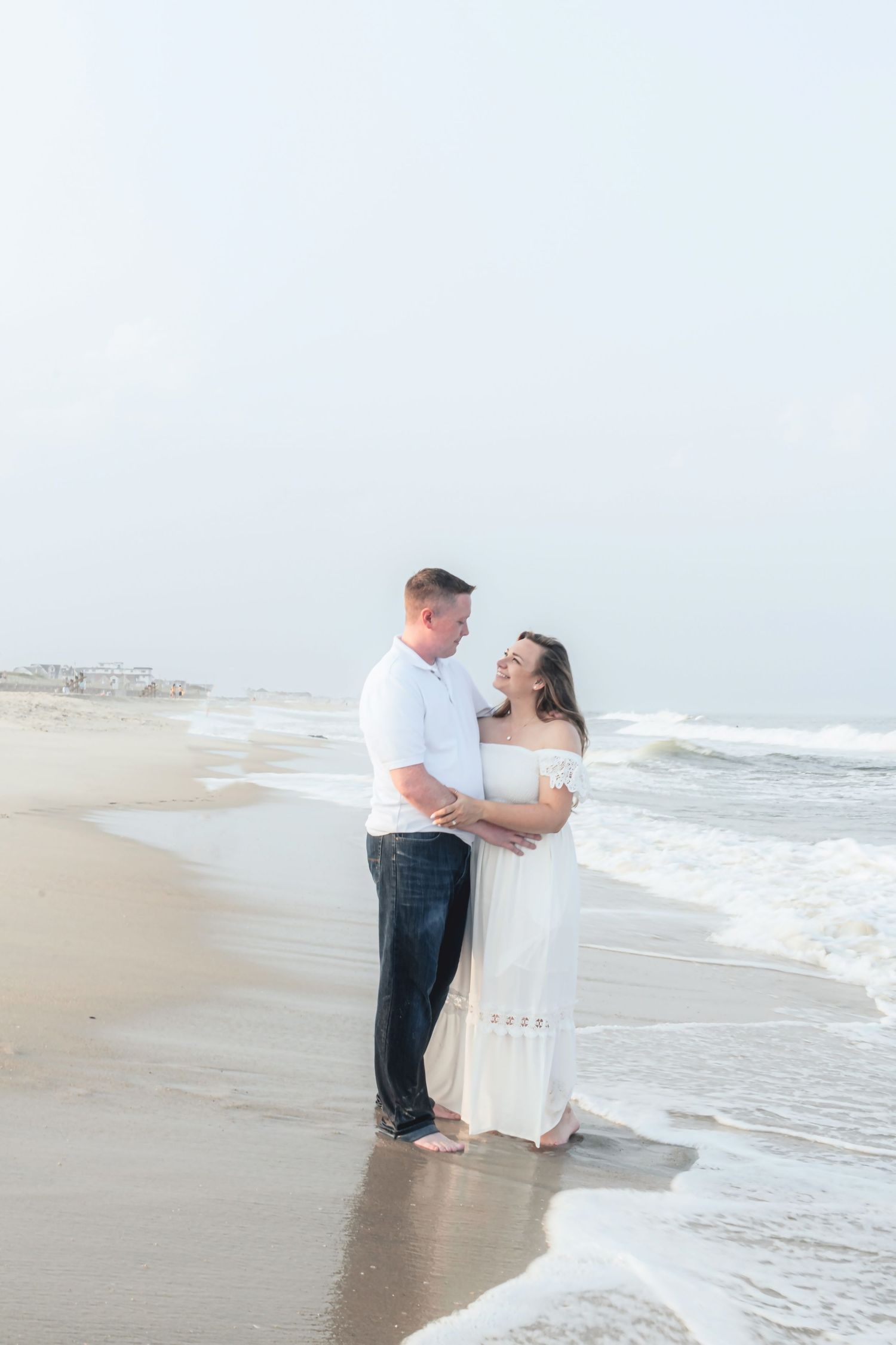 Couple embracing on a sandy beach in white outfits, smiling at each other with ocean waves in the background.