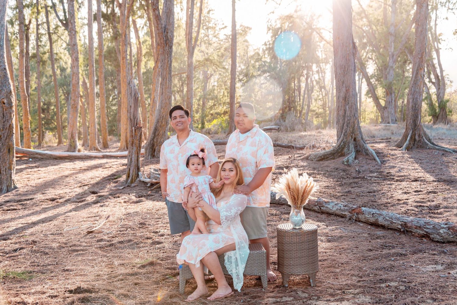 Family of four posing in a sunlit forest, wearing white outfits, with warm golden light filtering through tall trees.
