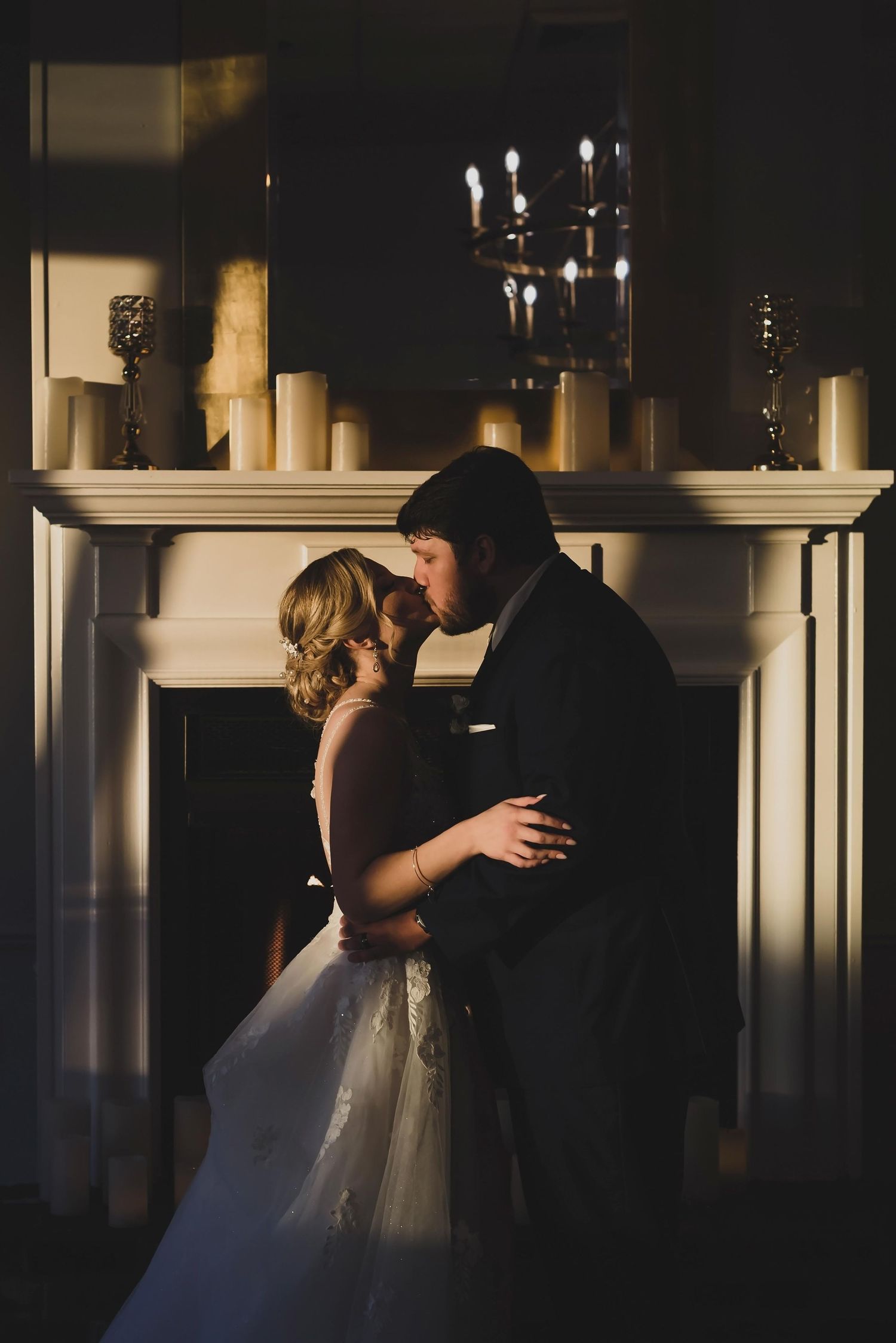 A romantic wedding couple embraces in front of a candlelit fireplace mantel creating a dramatic silhouetted scene.