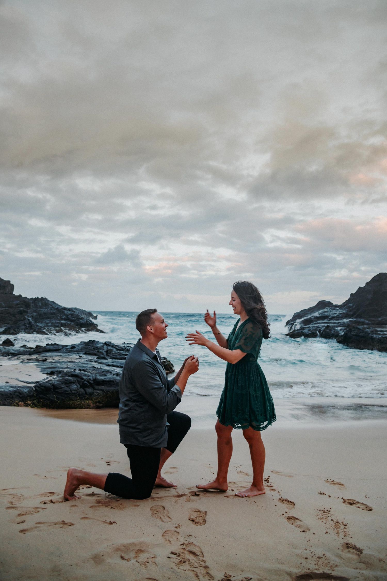 Couple shares a romantic proposal moment on a rocky beach during dramatic cloudy sunset.