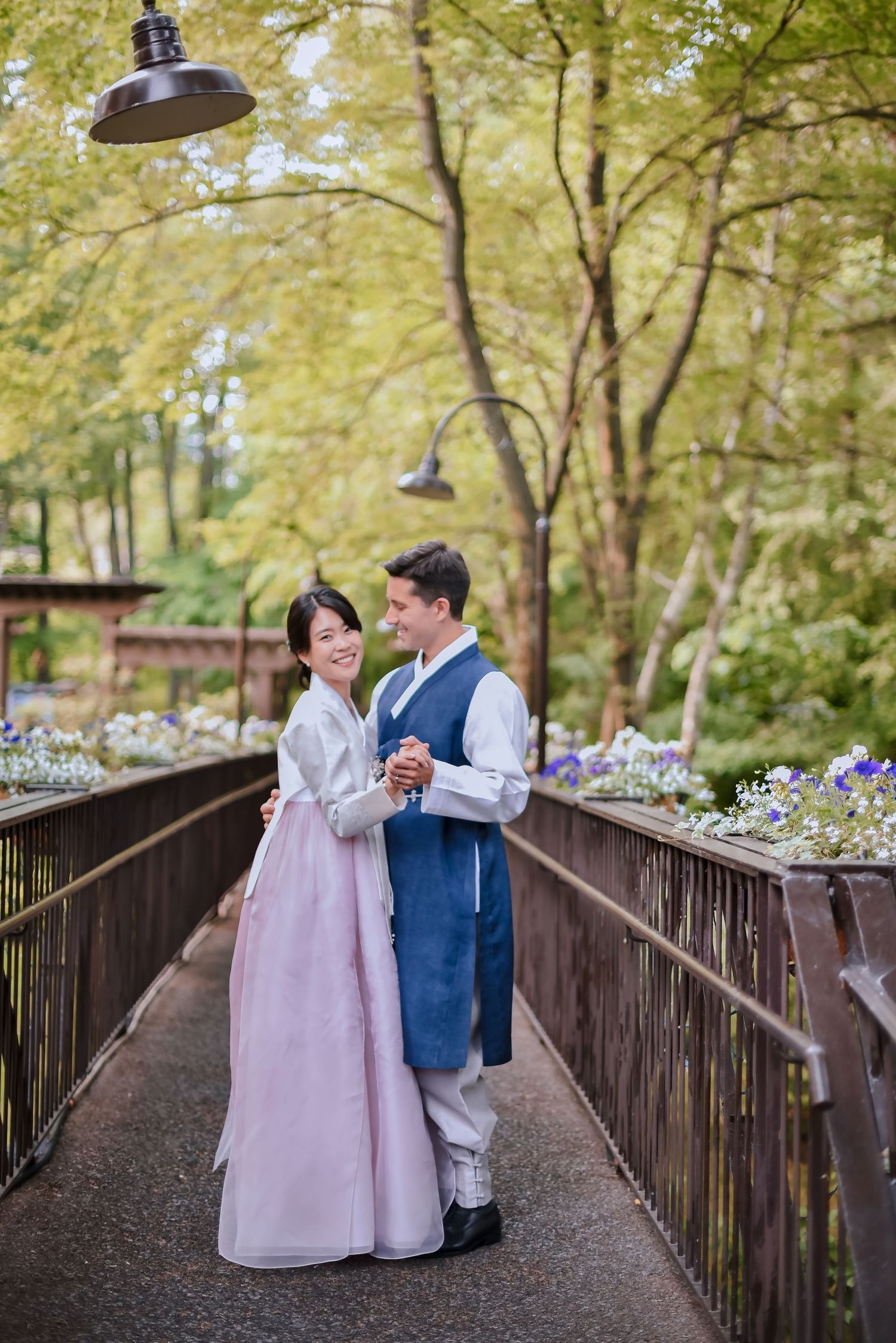 Two people in traditional Korean hanbok attire stand together on a wooden bridge path surrounded by autumn trees.