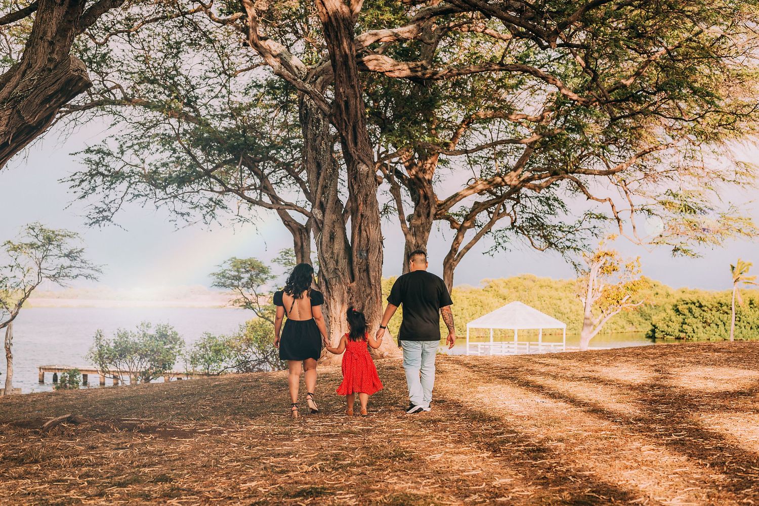 Family of three walking hand-in-hand toward ocean view under large tropical trees on sunny day.