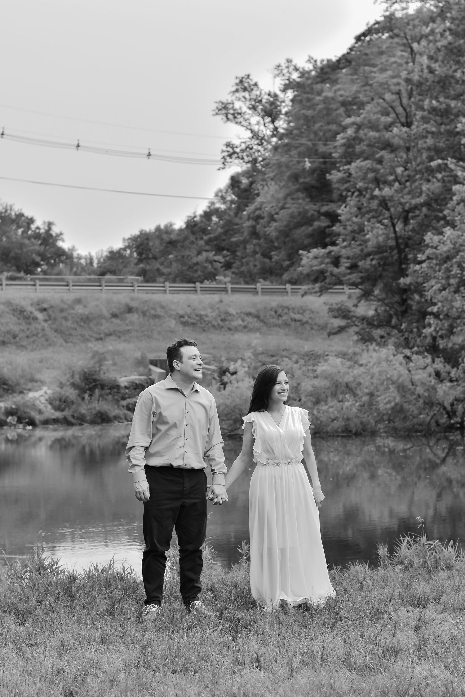 A couple in formal attire stands together by a serene lake surrounded by lush greenery in a romantic outdoor setting.