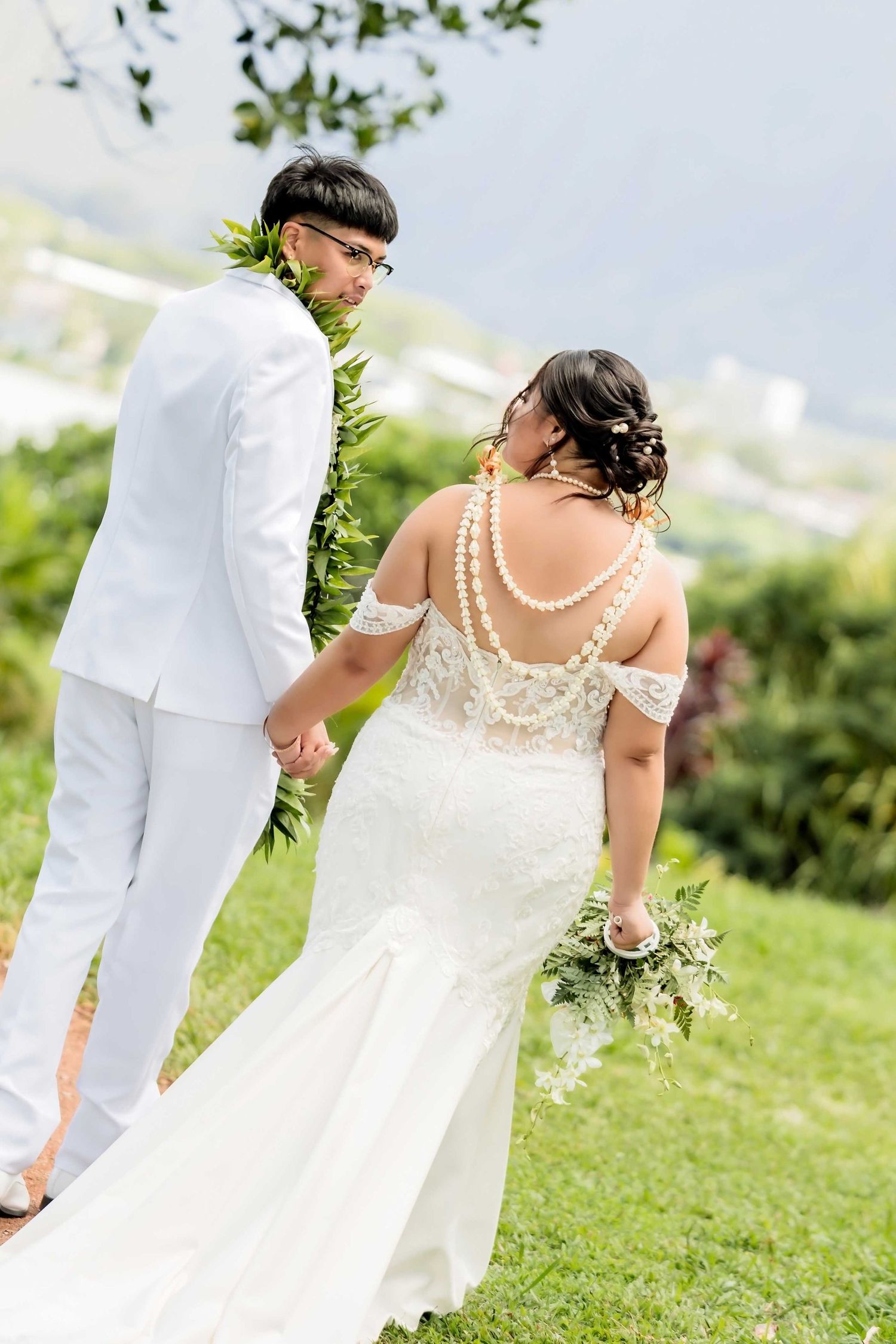Bride and groom holding hands outdoors in Hawaii, bride wearing lace gown with pearl back detail and groom in white suit.