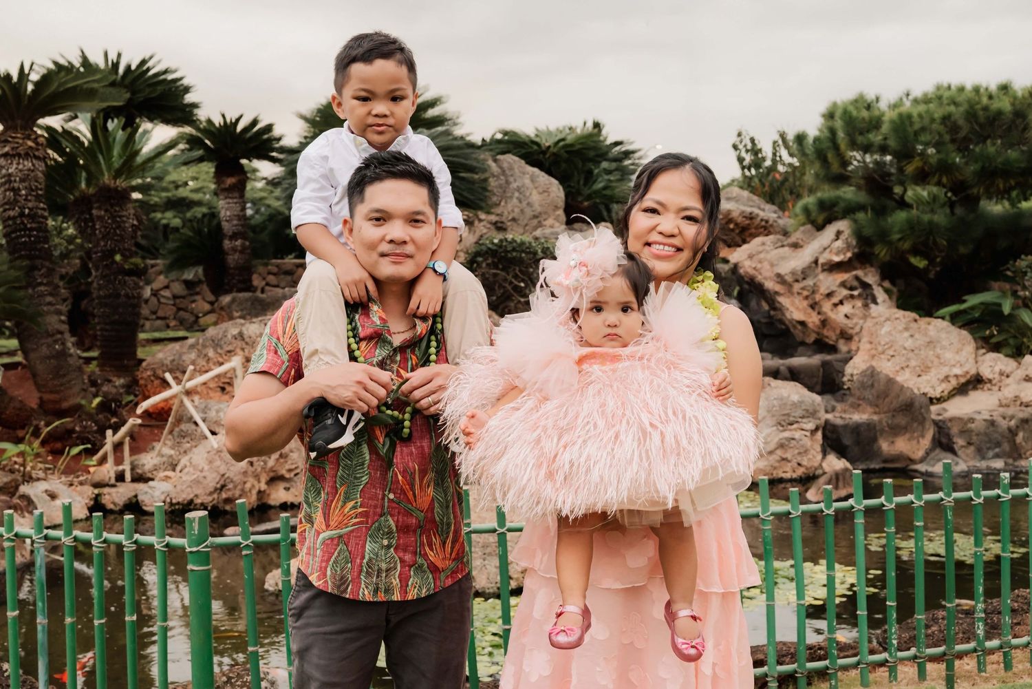 Family of four posing outdoors near a garden fountain, parents holding two young children dressed in colorful outfits.