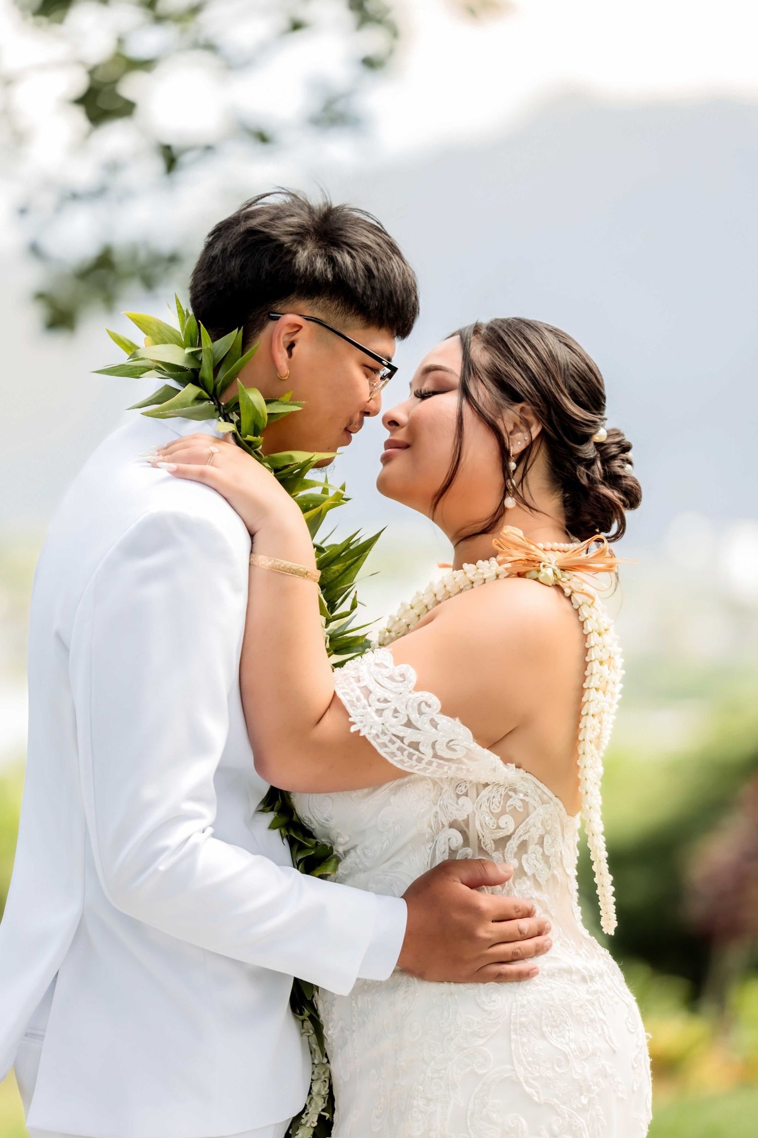 Bride and groom in white attire with Hawaiian leis share a romantic moment outdoors with mountain backdrop.