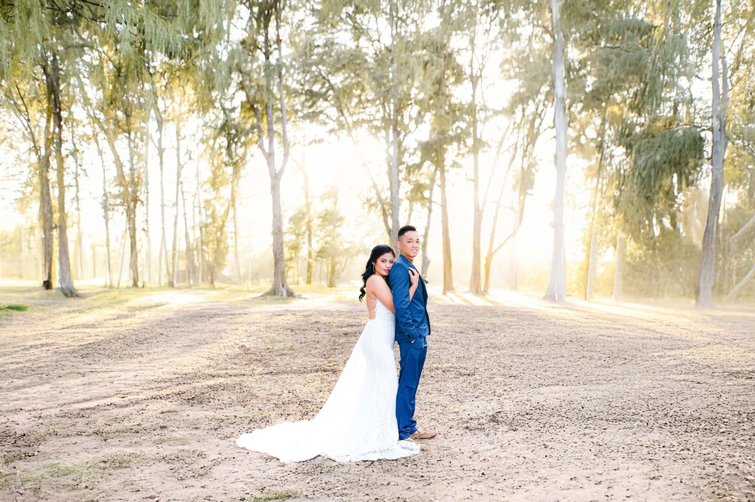 A couple in wedding attire walks through a sunlit wooded area during a romantic outdoor photo session.