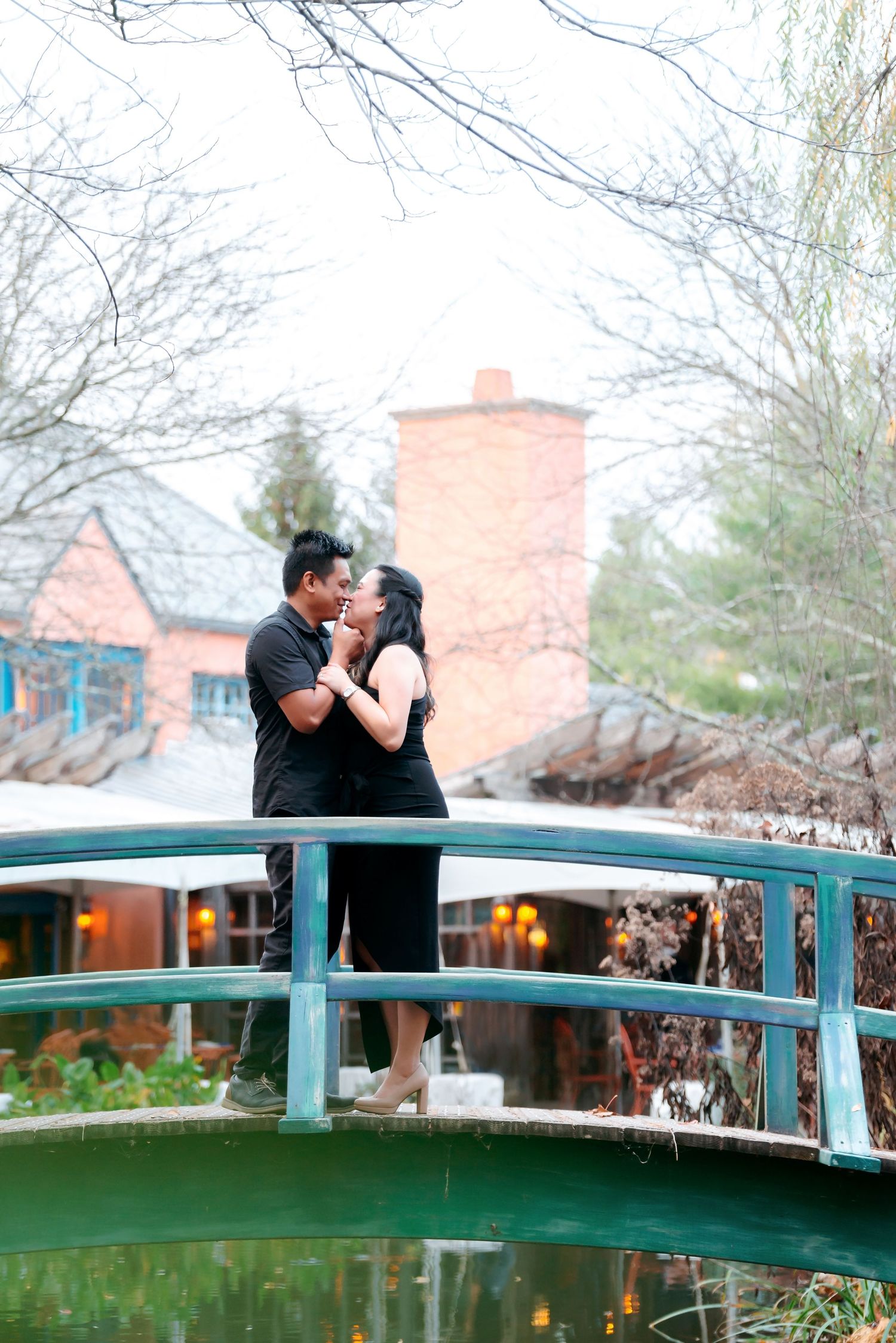 A couple shares a romantic moment on a green arched bridge in a garden setting with brick buildings in the background.