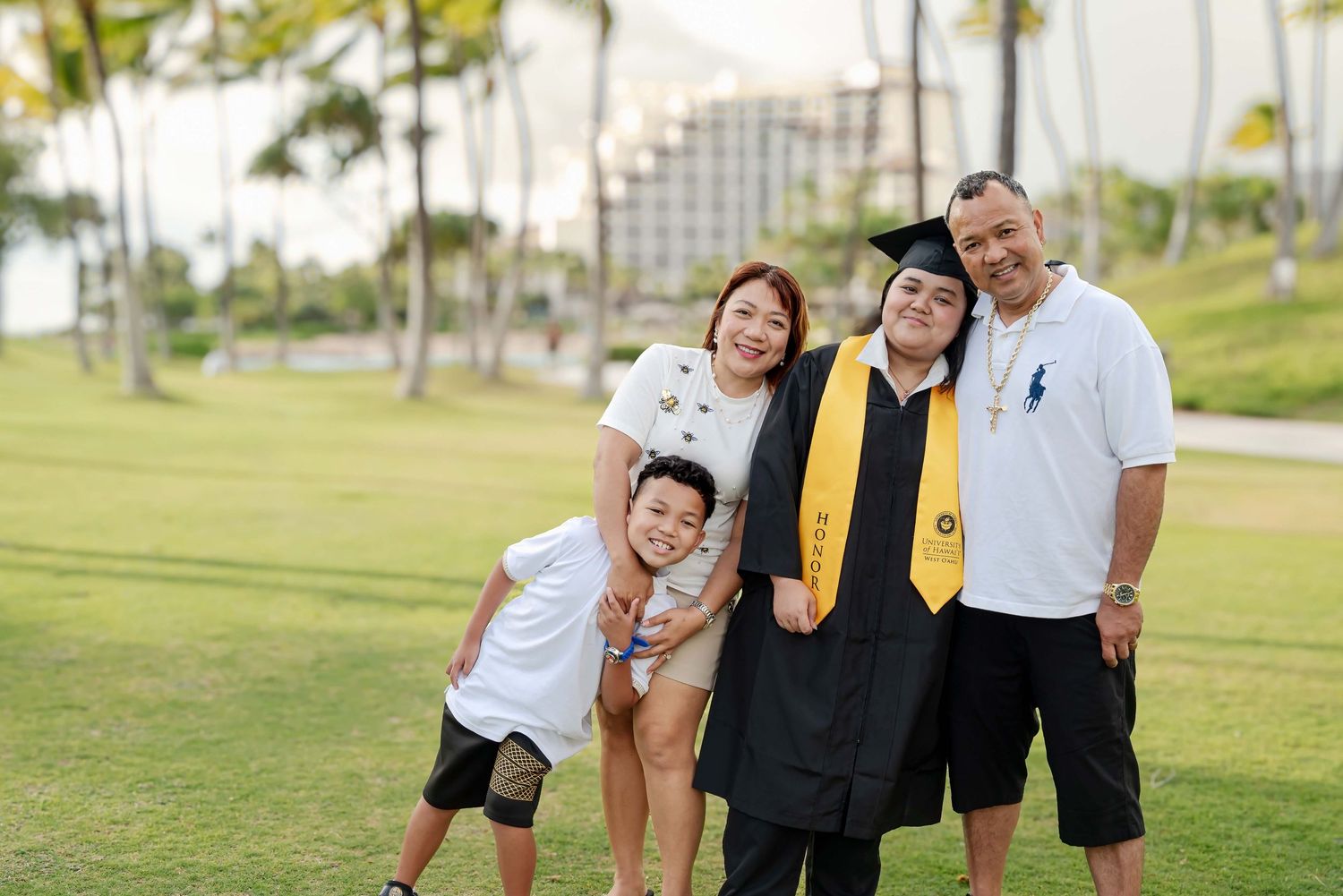 Family celebrating graduation outdoors on green lawn, graduate in cap and gown with yellow stole, palm trees and building behind.