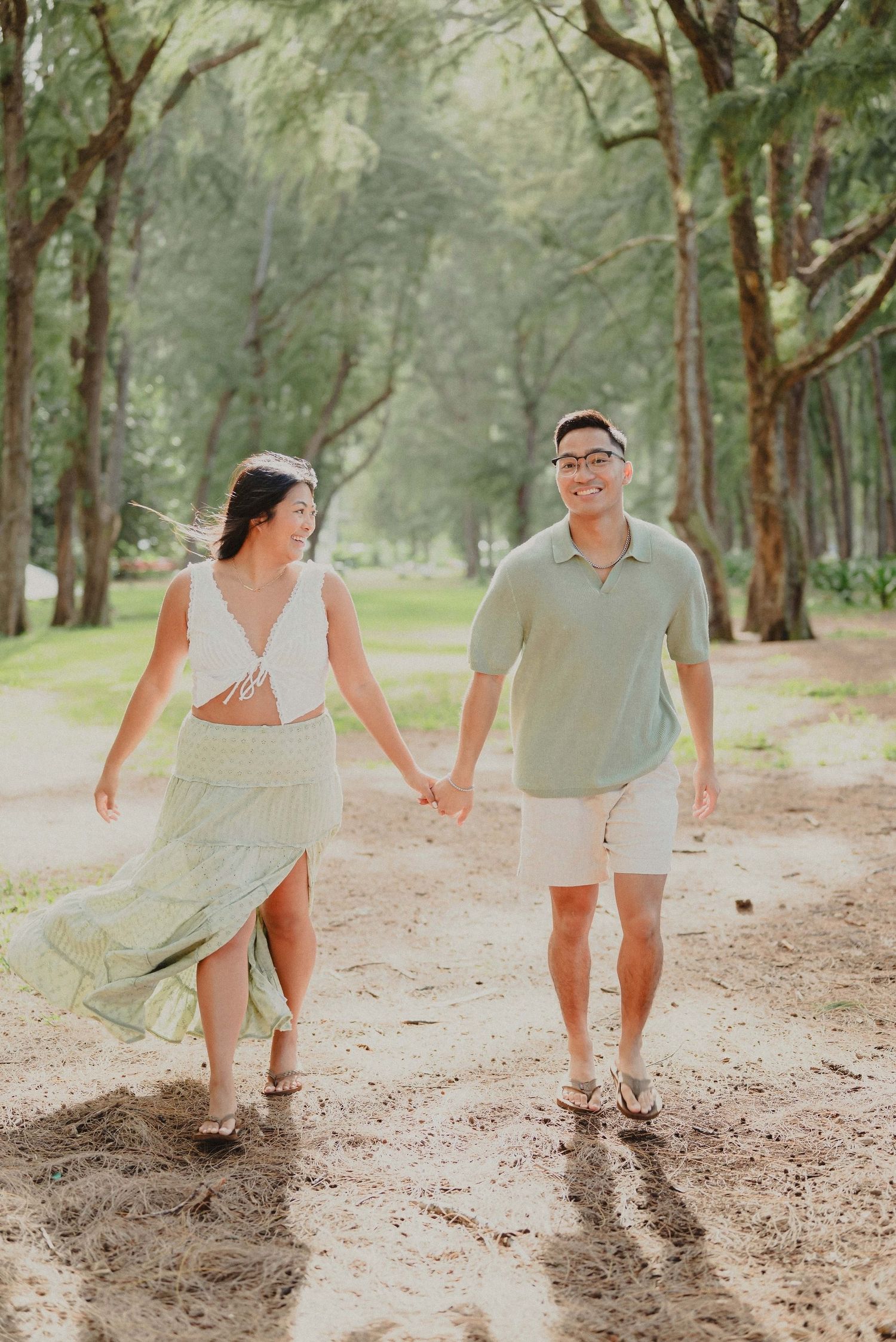 Couple holding hands walking on a sandy path through lush green trees in a tropical setting.